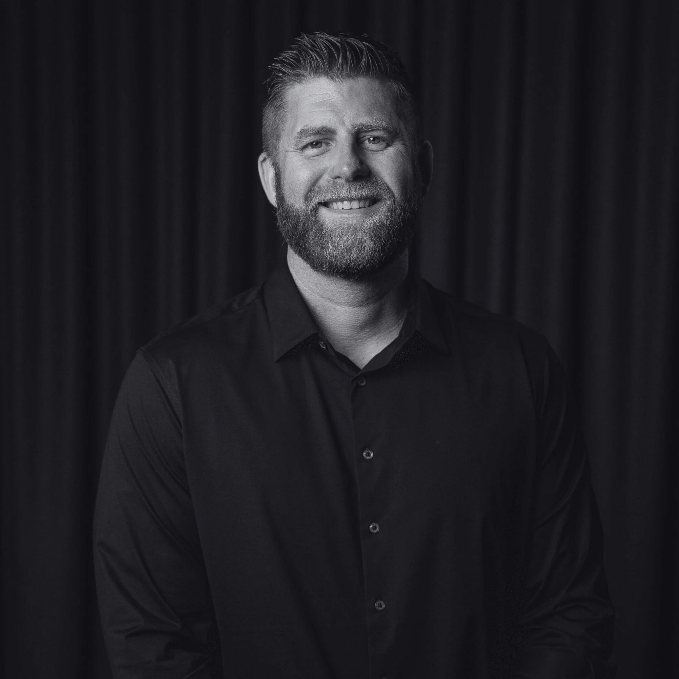 Black and white portrait of a smiling man with a beard, wearing a dark collared shirt, standing in front of a dark curtain background.
