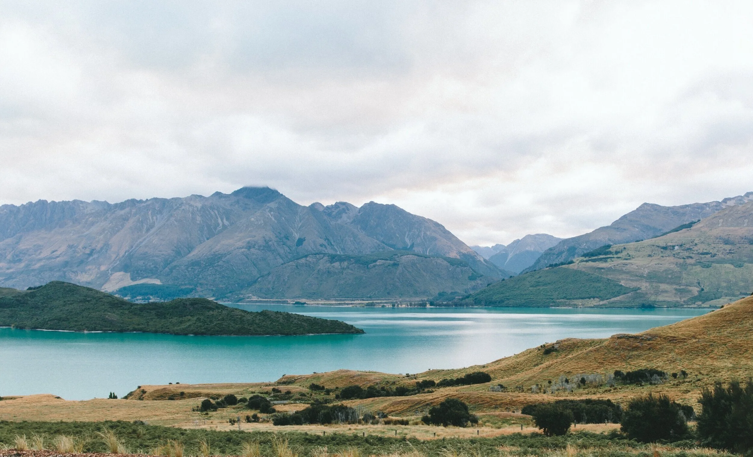 Scenic view of a large turquoise lake surrounded by rolling green hills and rugged mountains under cloudy sky.