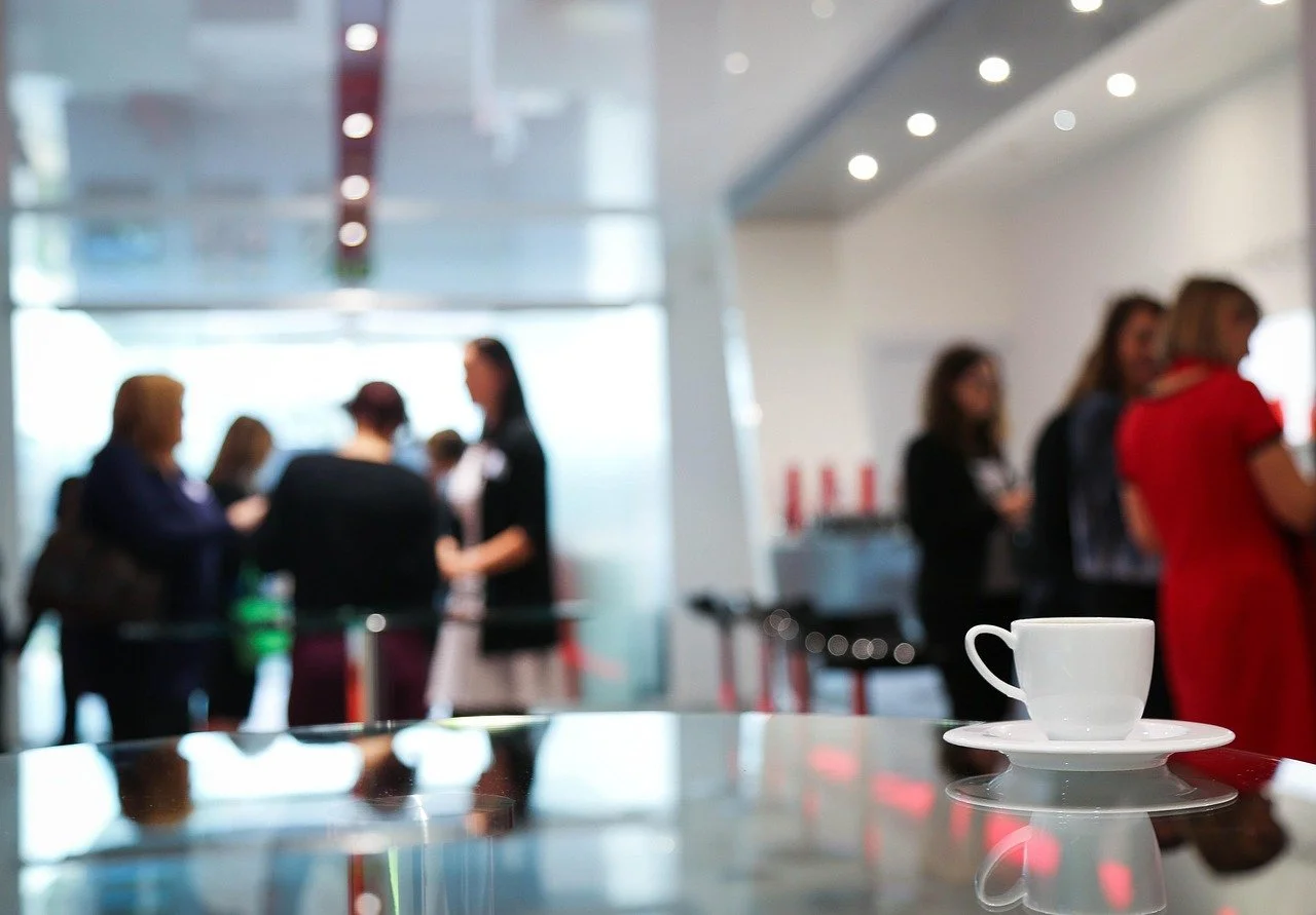 A white coffee cup with a matching saucer on a reflective surface in a busy cafe or waiting area, with blurred people in the background.