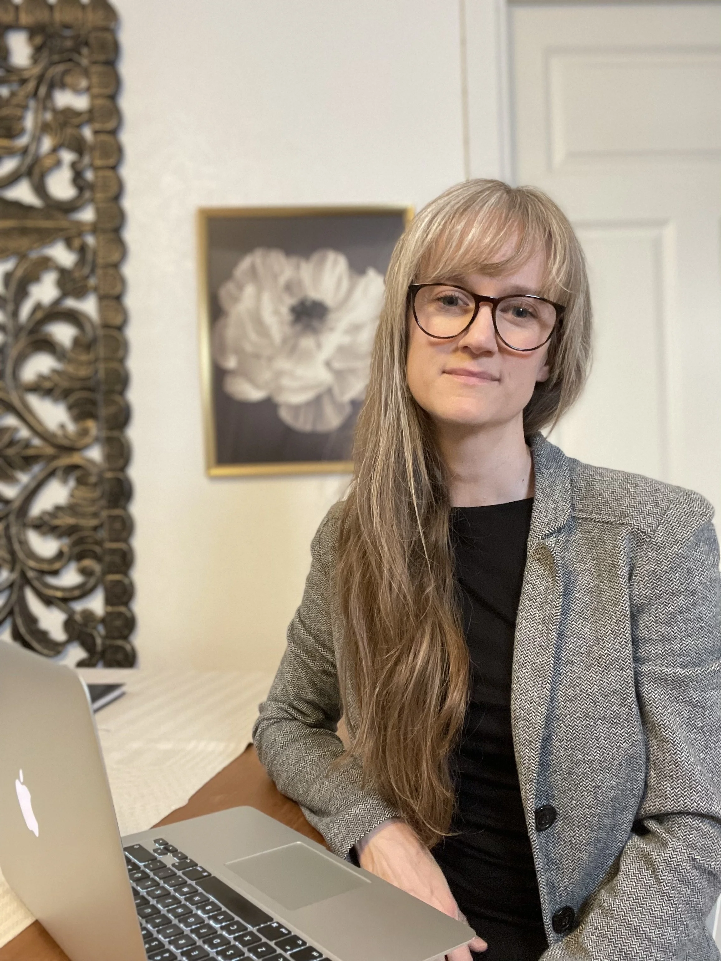 A woman with long hair, glasses, and a blazer sitting at a table with a MacBook laptop, in a room with a decorative wall and a framed flower picture in the background.