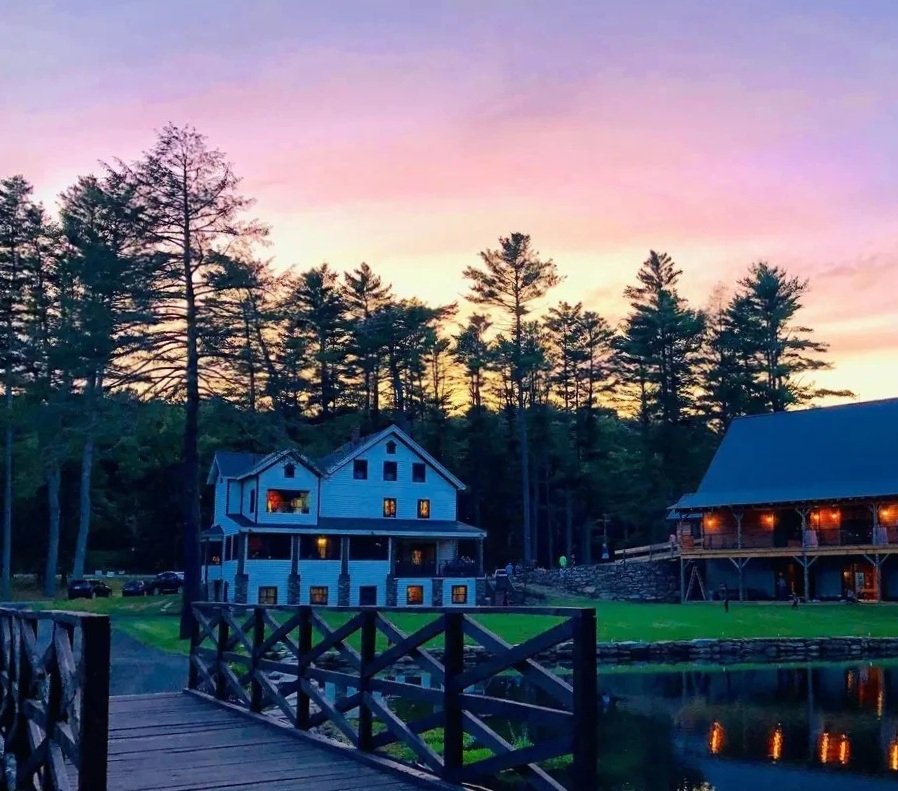 A scenic view of a pond with a wooden dock at sunset. In the background, there are trees and two large buildings with warm lights, one on the left and one on the right.