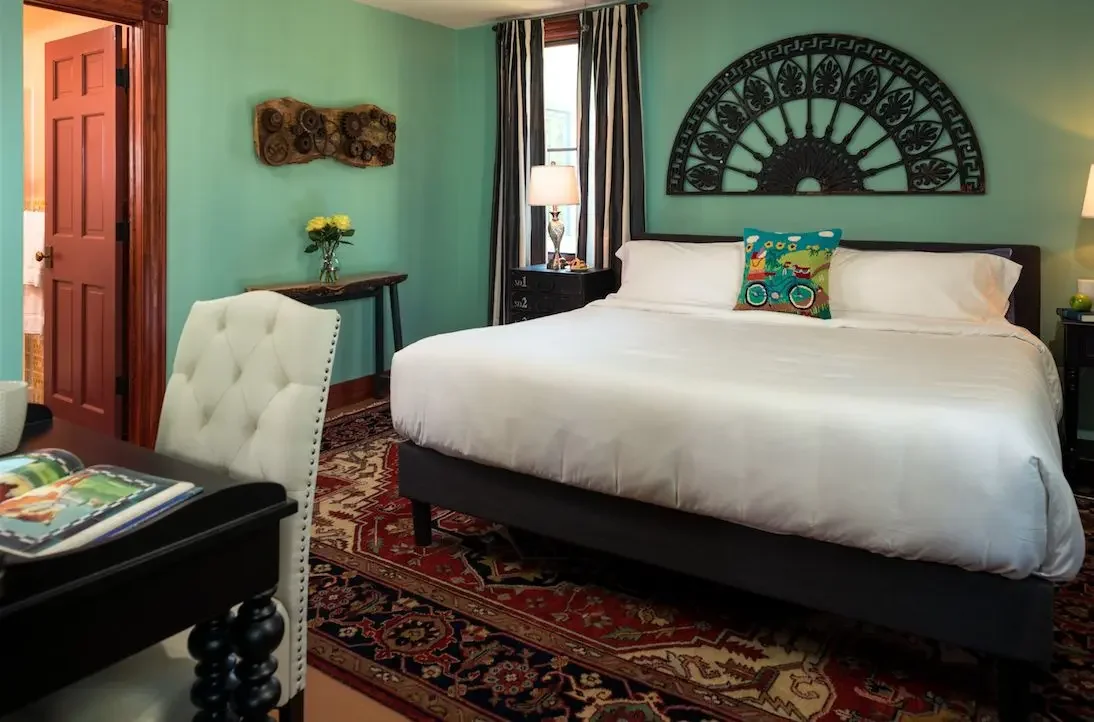 A bedroom with a large bed, white bedding, and a colorful pillow, wooden nightstands with lamps, a window with curtains, a green wall with art, a white chair, and a patterned rug.