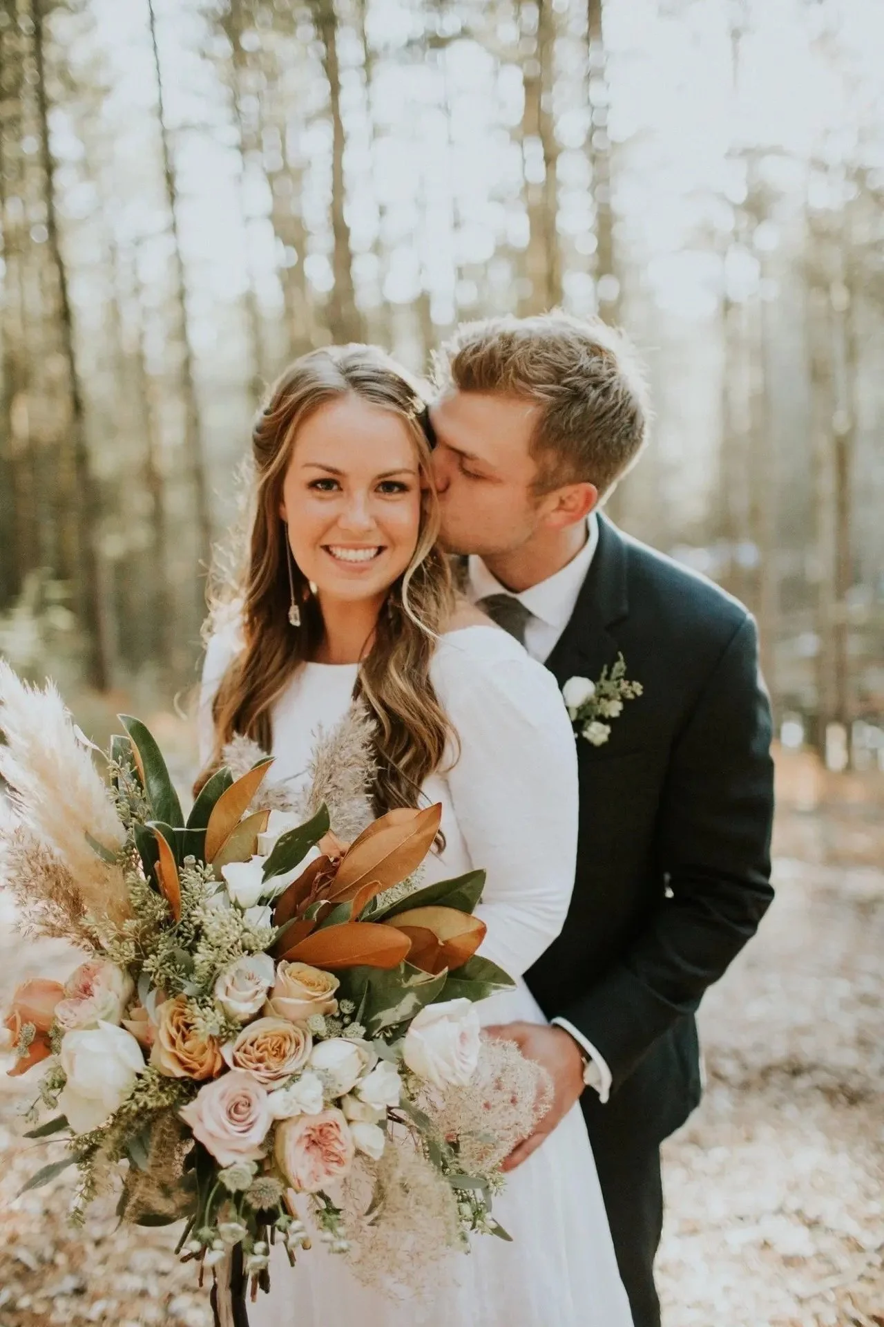 A bride holding a bouquet of flowers, smiling, while a groom kisses her on the cheek in a forest setting.