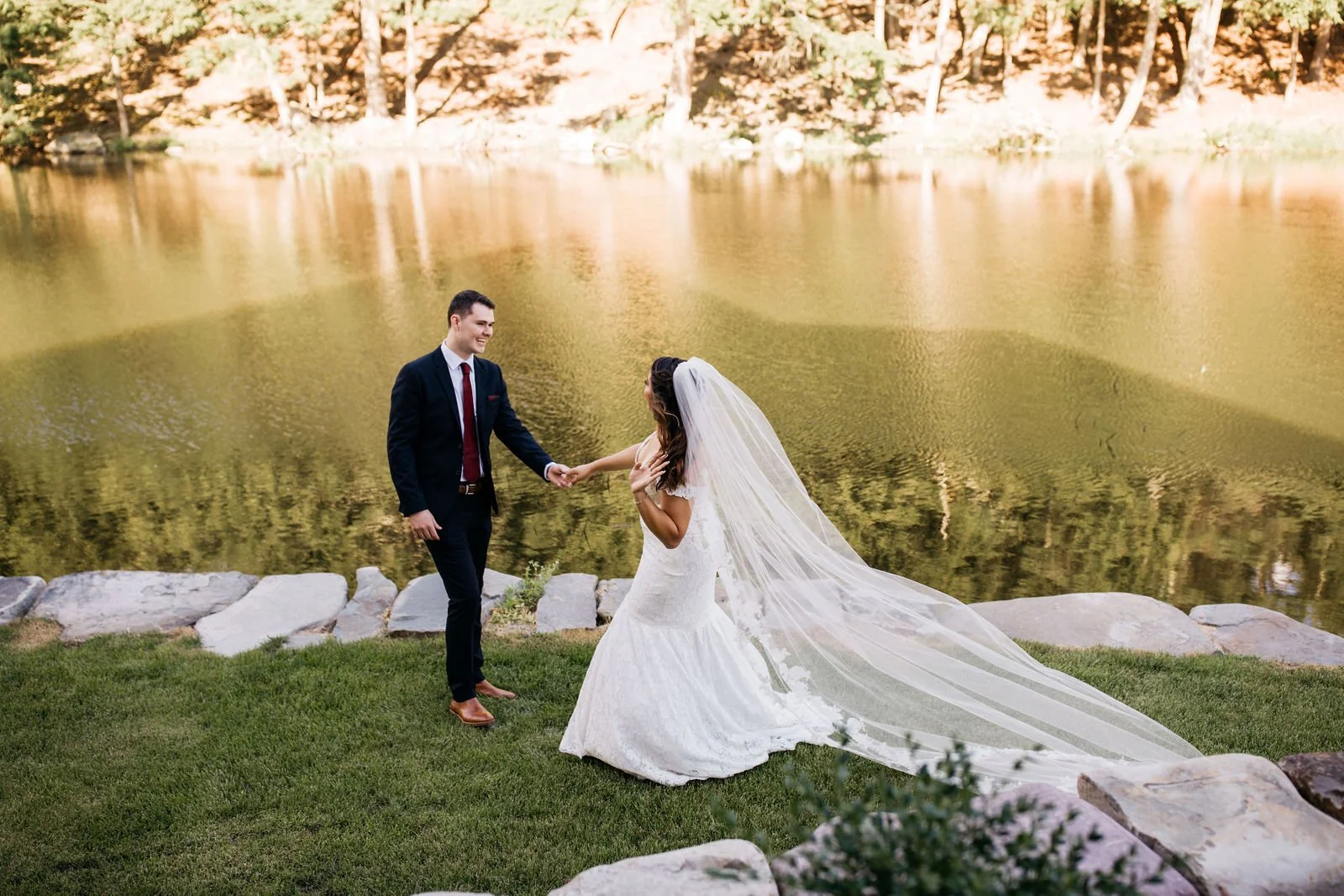 A bride and groom holding hands by a lake at sunset, with trees in the background.