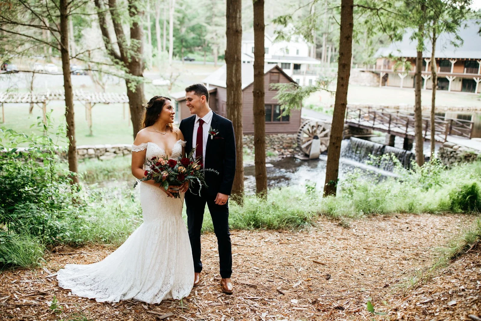 A bride and groom standing together outdoors in a wooded area, smiling at each other, with a small river and rustic buildings in the background.
