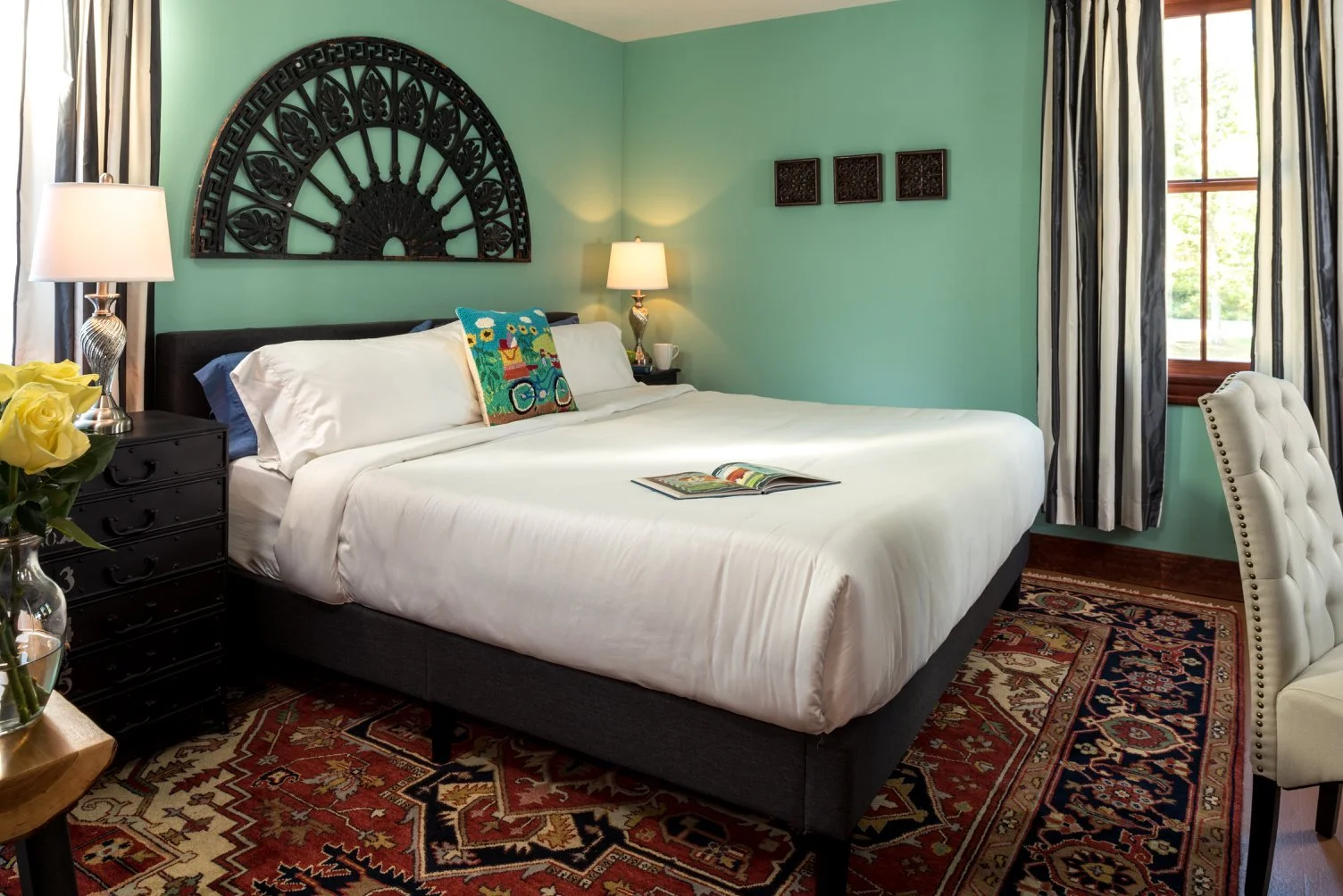 A cozy bedroom with a large bed, white bedding, colorful pillows, black bedside tables with lamps, a window with curtains, and decorative wall art.