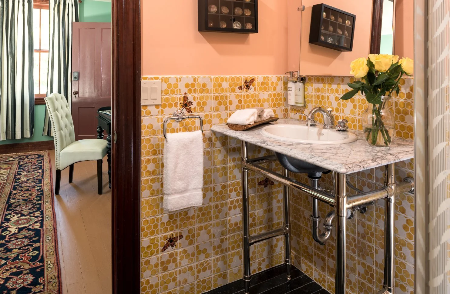 A bathroom with a marble sink, yellow honeycomb tile backsplash with butterfly decorations, a vase of yellow roses, and hanging black shelves. The adjacent room has a chair with a white upholstered back and a green curtain.