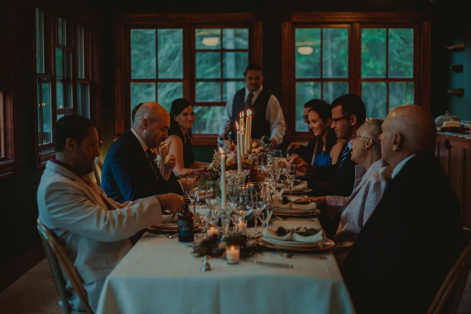 People sitting around a decorated dinner table with lit candles, many wearing formal attire, in a warmly lit room with large windows showing an outdoor scene.