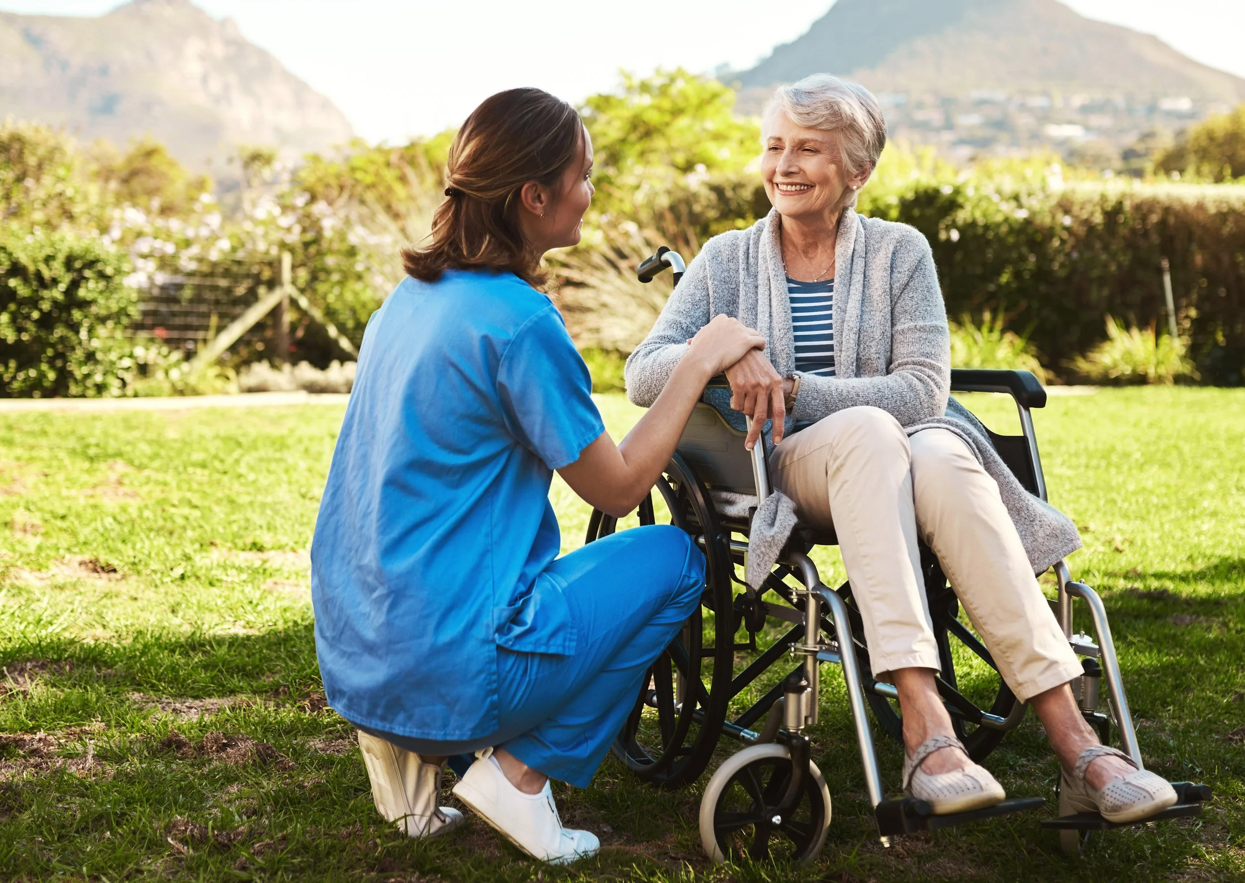 A nurse talking to a smiling elderly woman in a wheelchair outdoors on a sunny day, with greenery and mountains in the background.