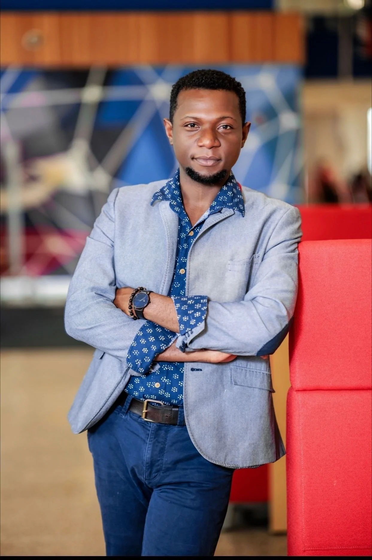 A young man with short curly hair and a goatee, wearing a gray blazer over a blue patterned shirt, standing with arms crossed next to a red chair inside a modern indoor setting with a colorful abstract background.