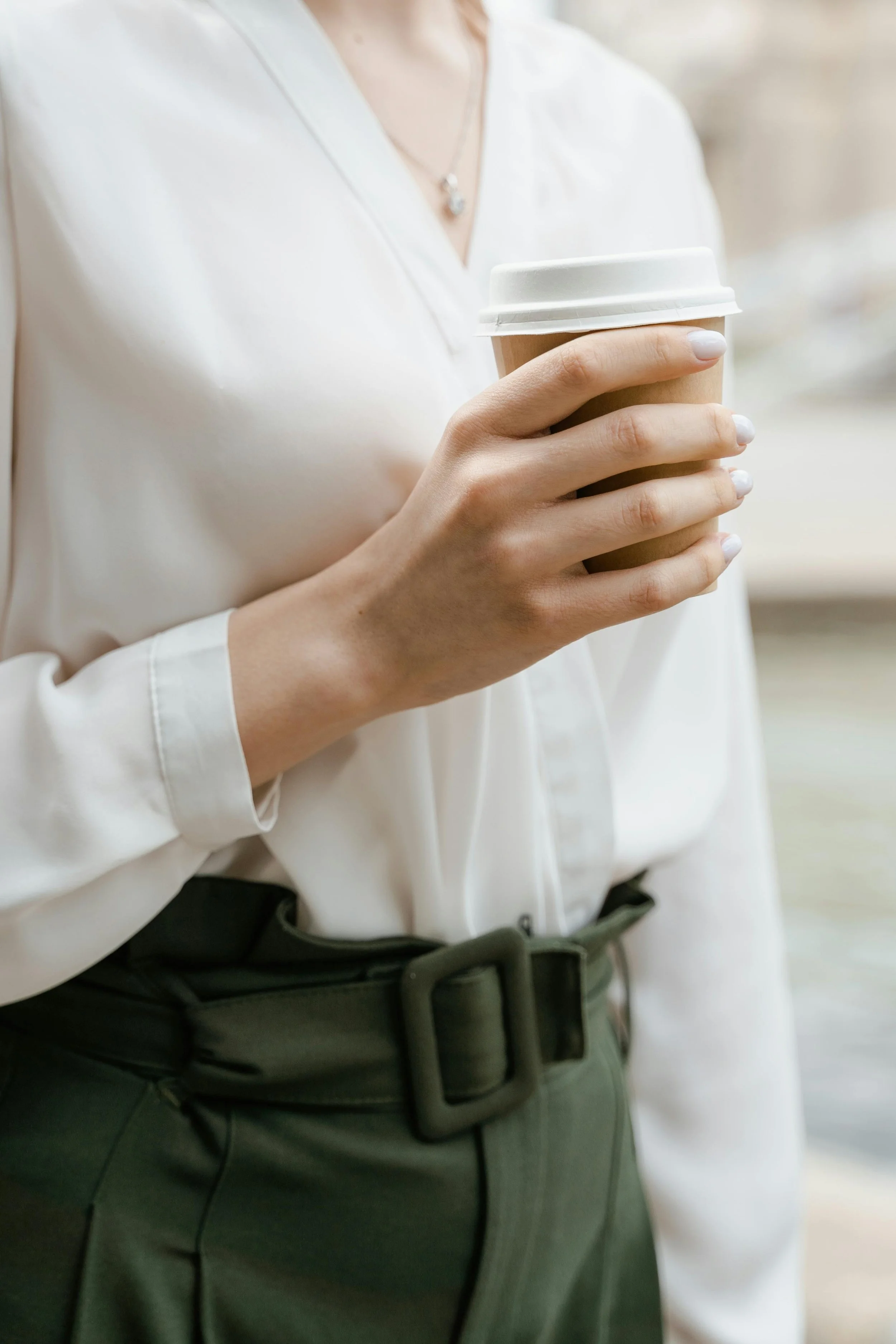 Close-up of a woman holding a disposable coffee cup with a white lid, wearing a white blouse and a dark green belt.