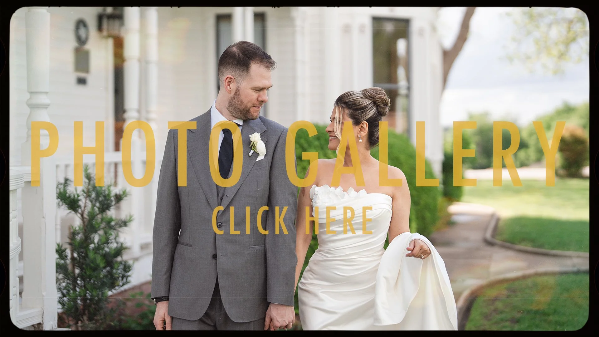 A bride and groom in wedding attire holding hands, smiling at each other outdoors in front of a white house with greenery