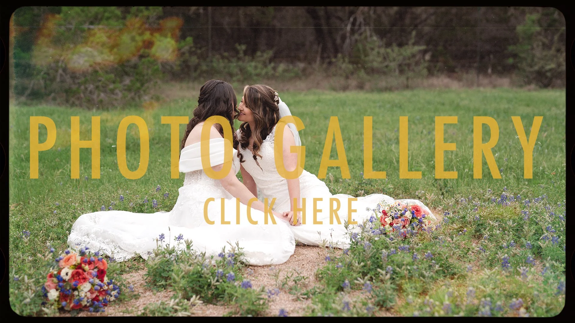 Two women in wedding dresses sitting on grass in a field, touching foreheads and holding hands, with a bouquet of flowers nearby, and trees in the background.