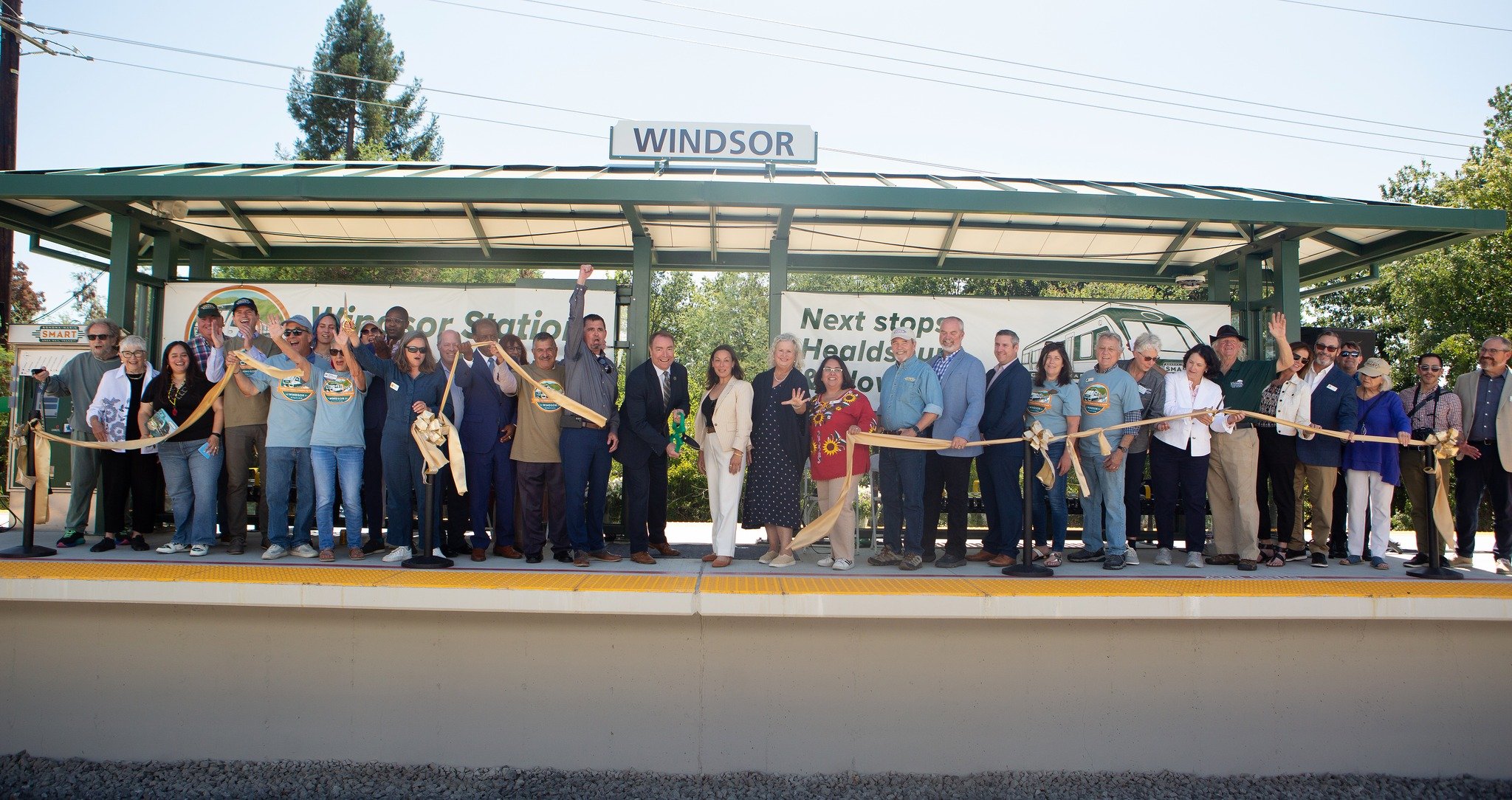 A group of people attending a ribbon-cutting ceremony at Windsor station, holding a ribbon and posing for a photo under a sign that says "Windsor."