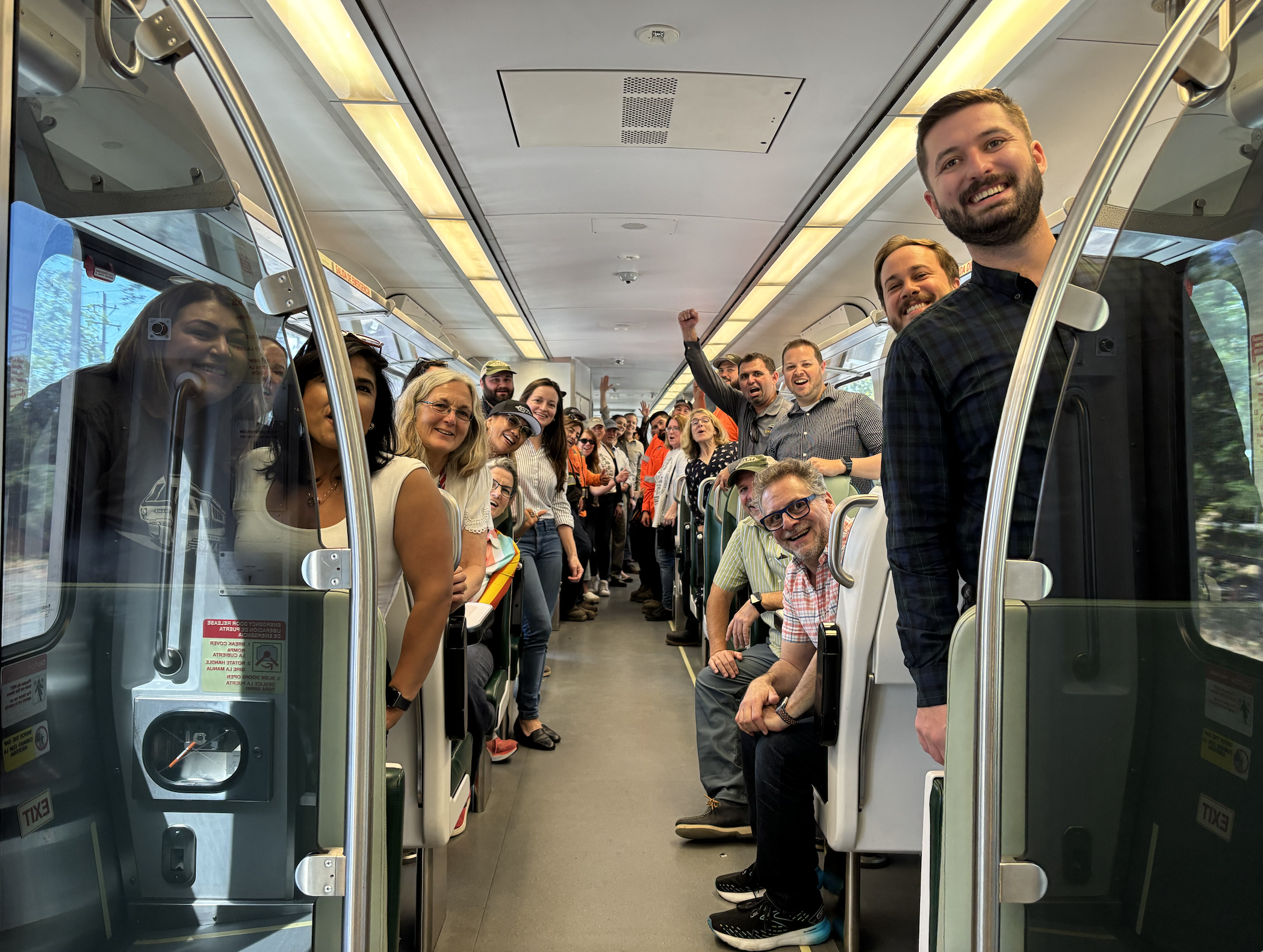Group of people smiling and looking at the camera inside a bus, some standing and some sitting, with a few raising their fists in celebration.