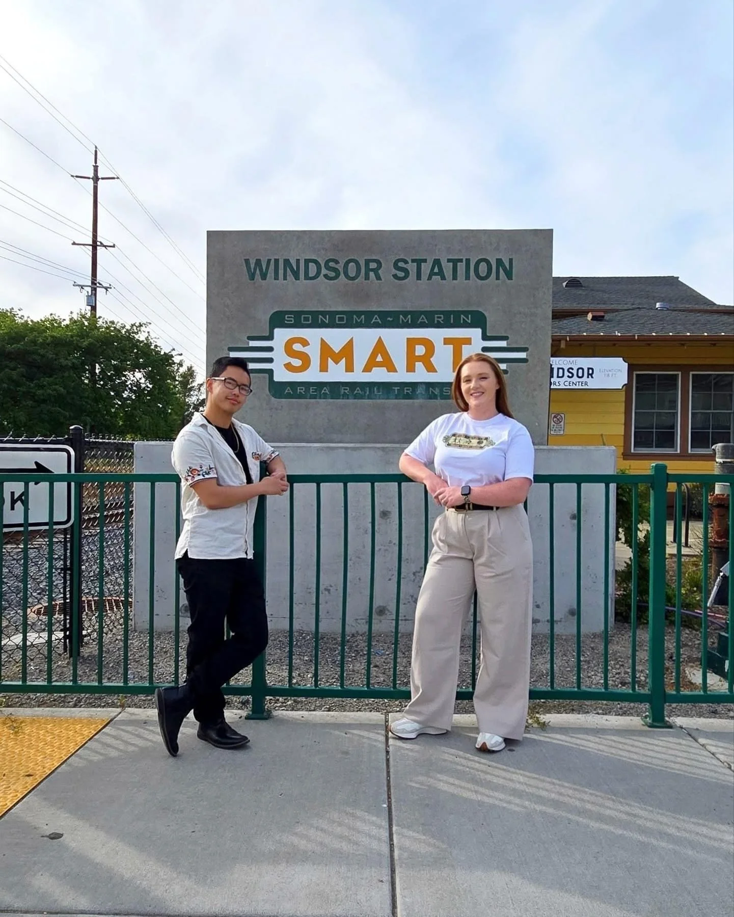 Two people standing in front of a sign at Windsor Station, smiling. The sign reads 'Windsor Station Sonoma Marin SMART Area Rail Transit'.