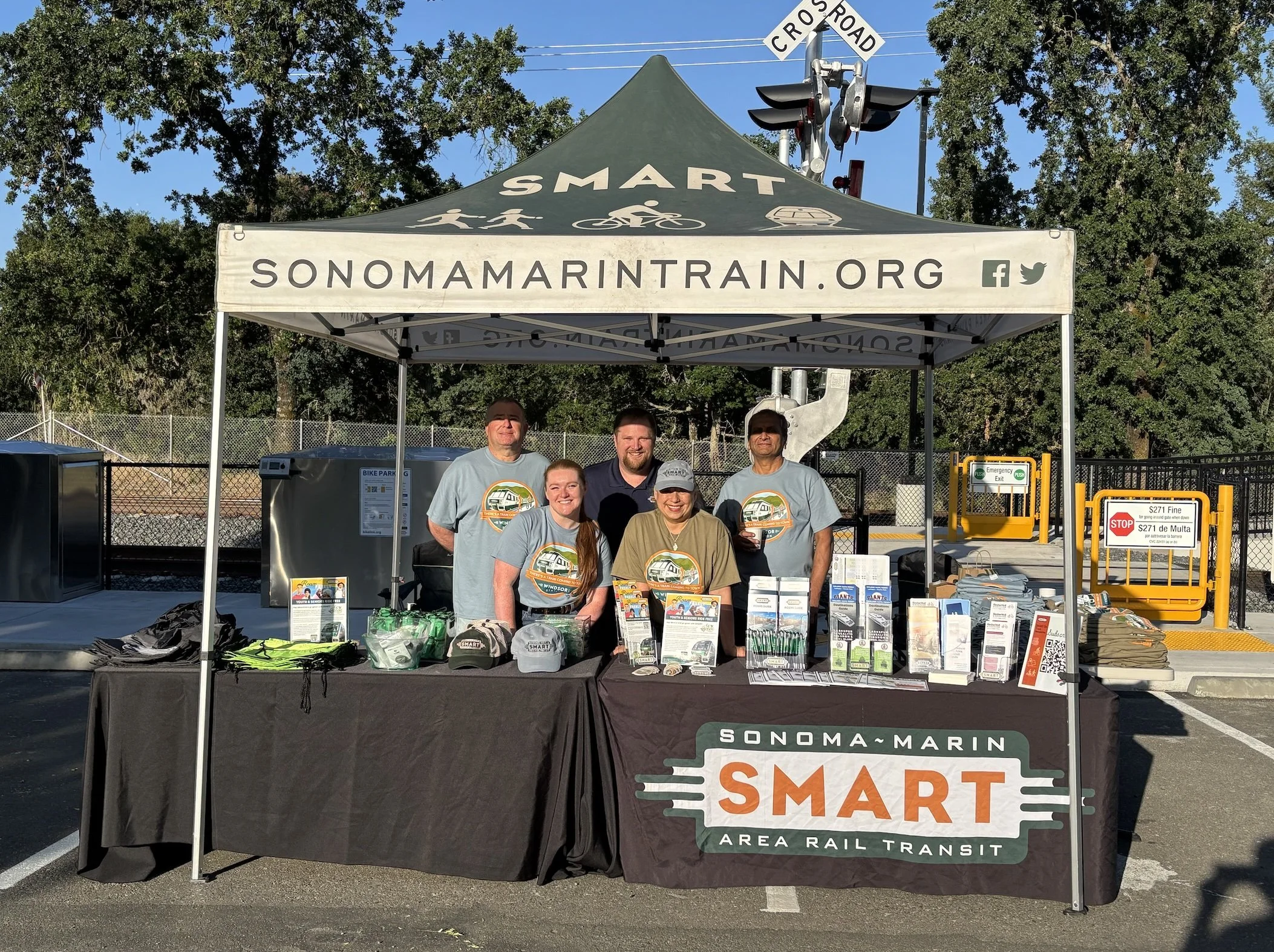 Group of five people standing behind a table under a tent promoting Sonoma-Marin Area Rail Transit. The tent has the website sonomamarintrain.org and logos on it, with brochures, hats, and promotional items on the table.