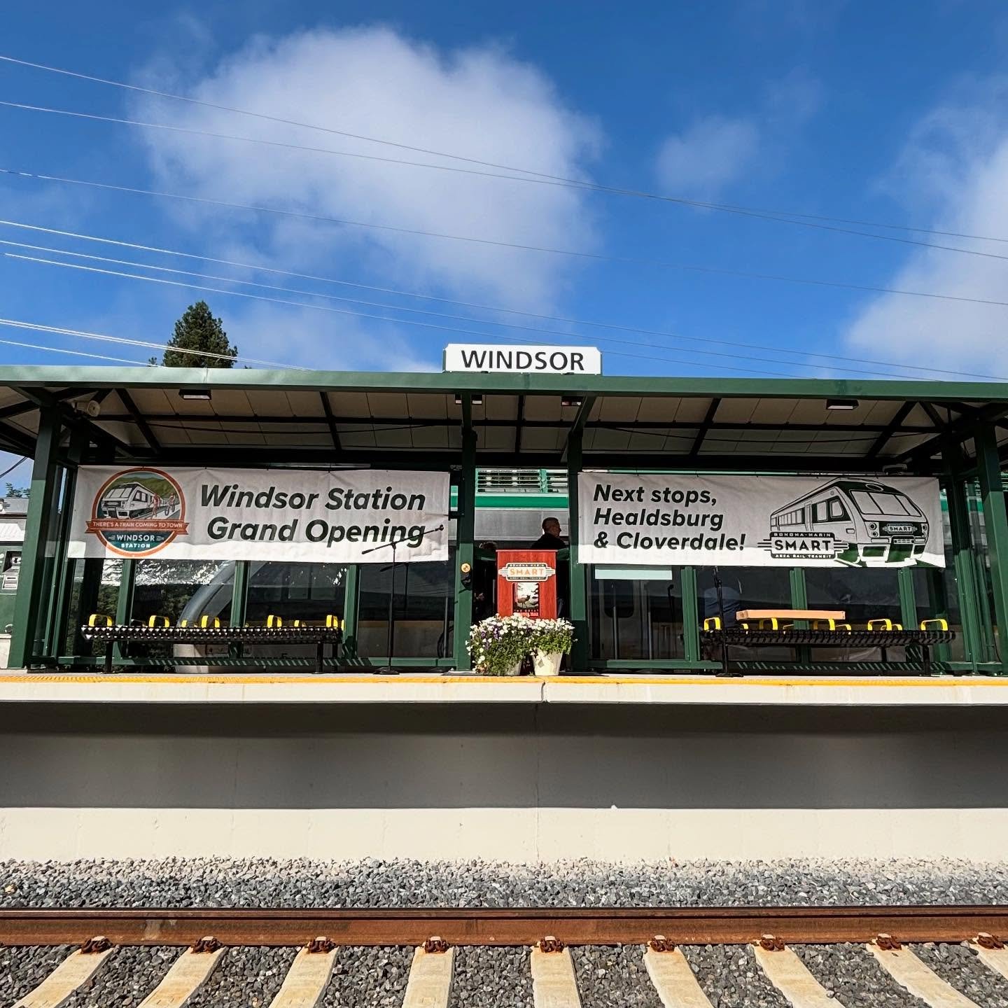 Windsor train station platform with banners announcing grand opening and next stops at Healdsburg and Cloverdale, with a train visible behind the banners, under a blue sky with clouds.