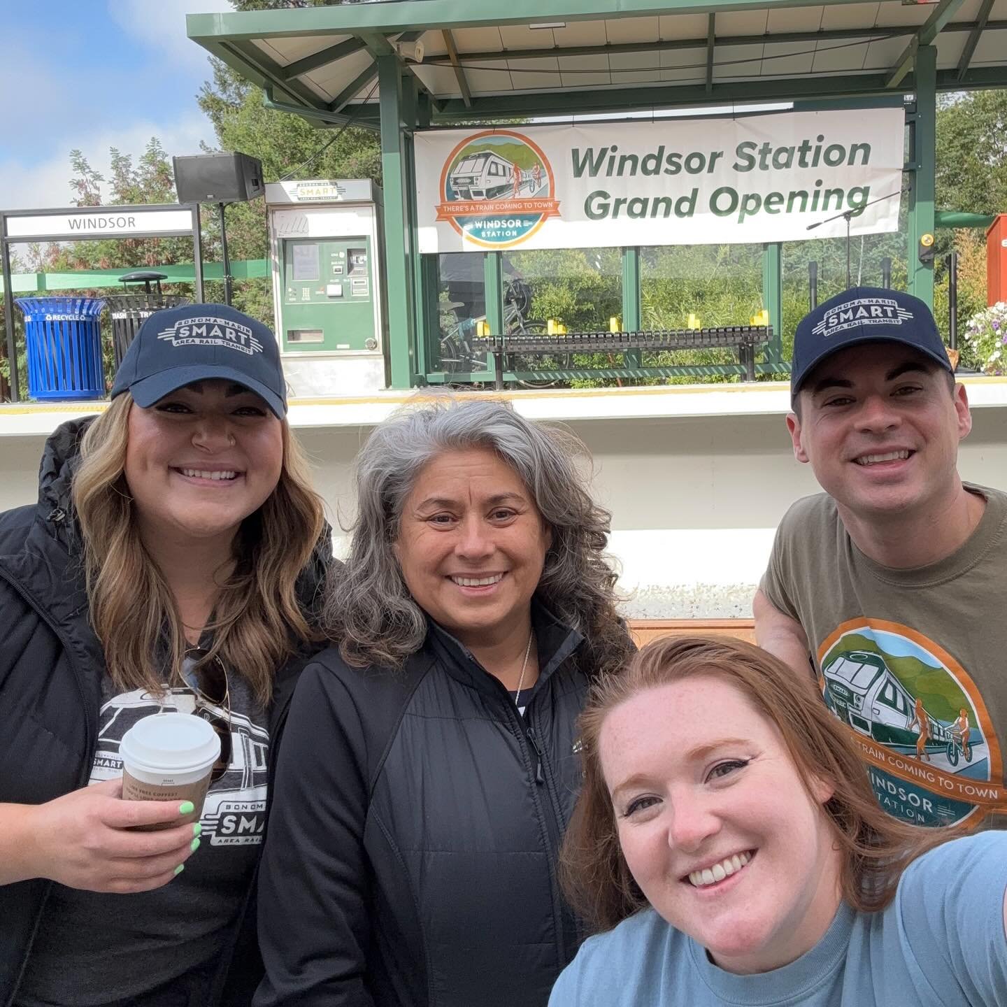 Group of four smiling people taking a selfie in front of Windsor Station with a banner that reads 'Windsor Station Grand Opening' in the background.