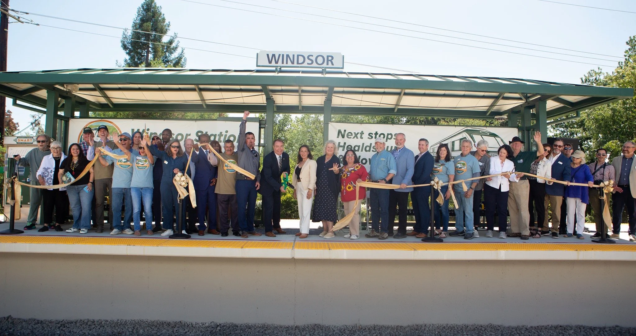 A large group of people participating in a ribbon-cutting ceremony at a train station, with the sign 'WINDSOR' above them. The group is standing on a platform, some holding a gold ribbon, with banners and trees in the background.