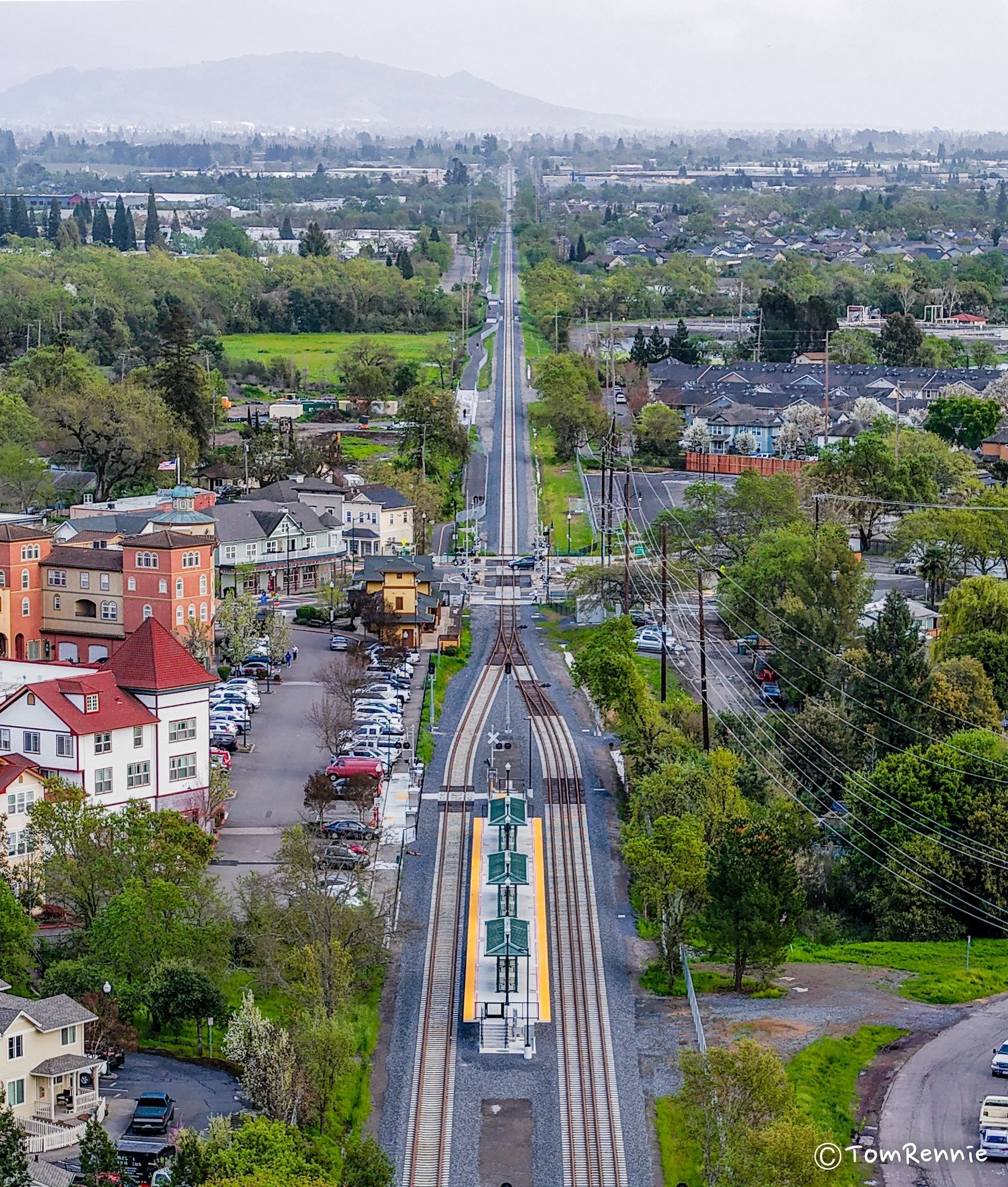Aerial view of a small town with a train station, train tracks, residential houses, and green fields extending towards distant mountains.