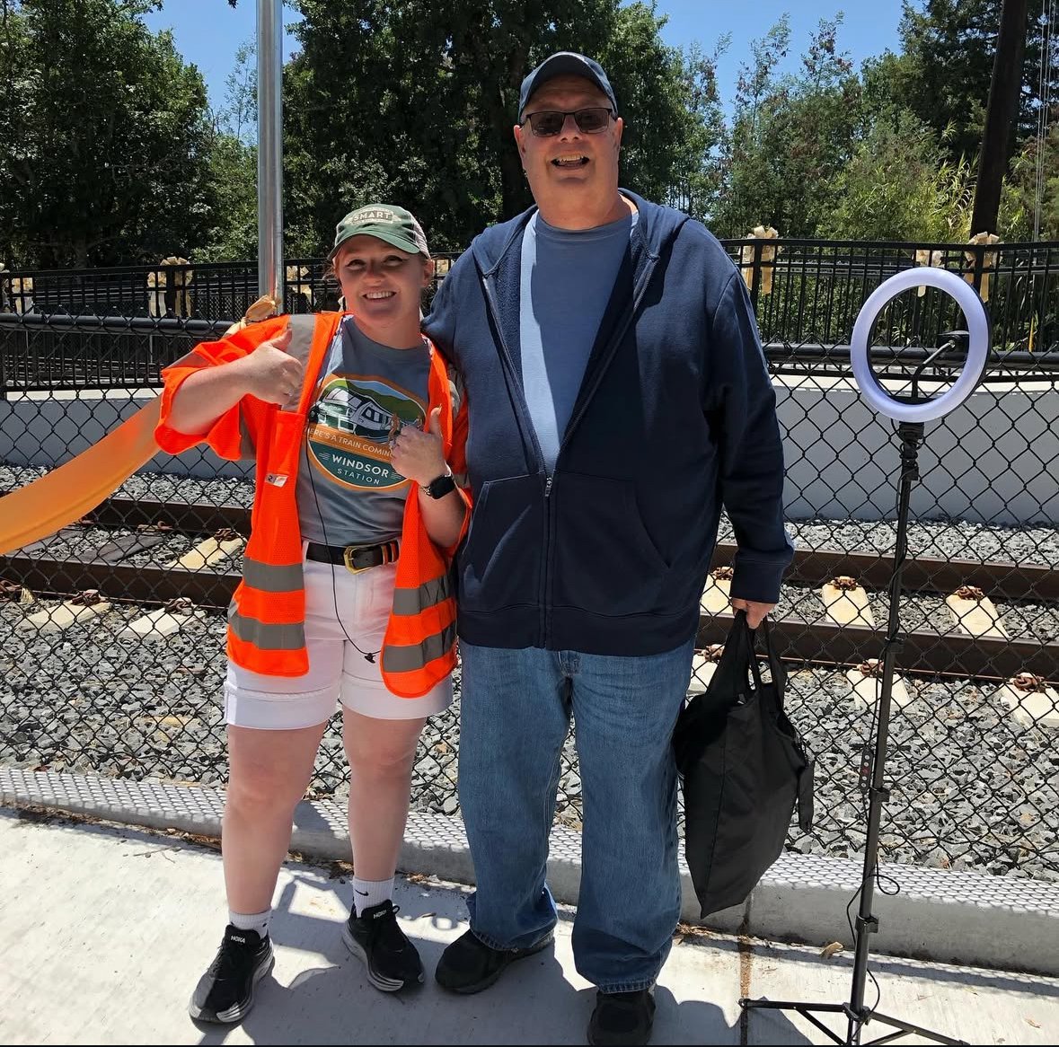 Two people standing in front of a train track fence outdoors, smiling at the camera. The person on the left is a young woman wearing a gray shirt, white shorts, black sneakers, and an orange safety vest, giving a thumbs-up. The person on the right is