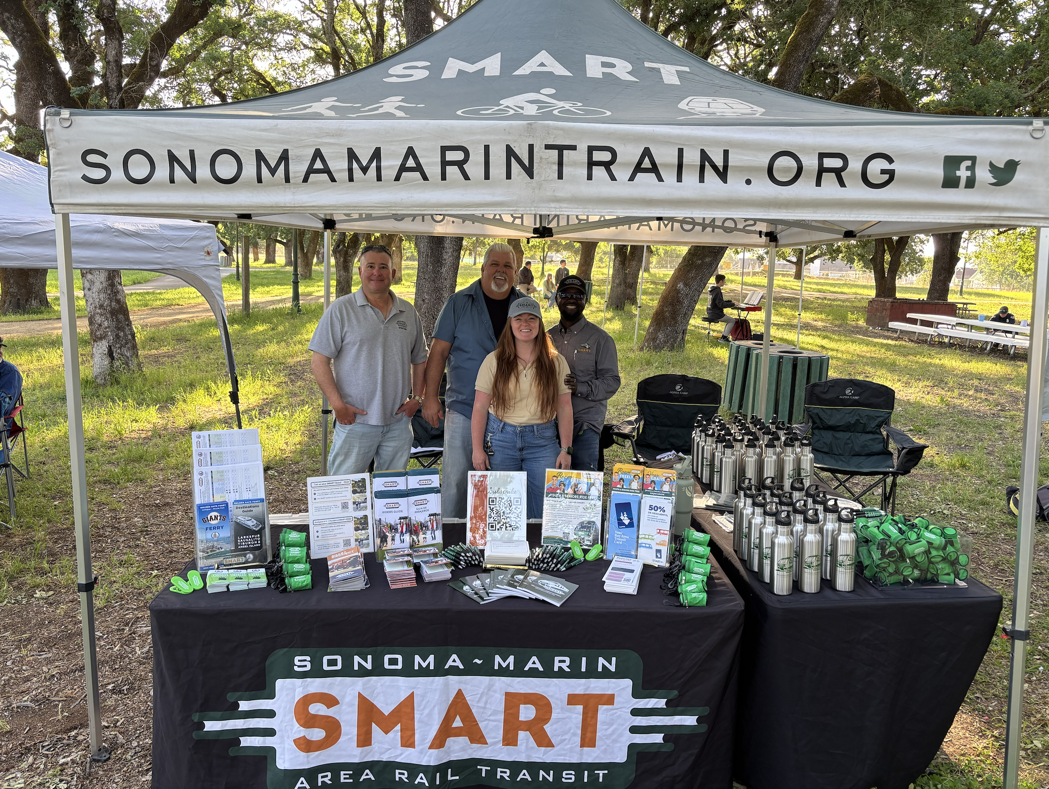 Four people standing behind a table under a canopy at an outdoor event, promoting Sonoma-Marin SMART Area Rail Transit, with informational materials, water bottles, and promotional items on the table. Trees and park benches are visible in the background.