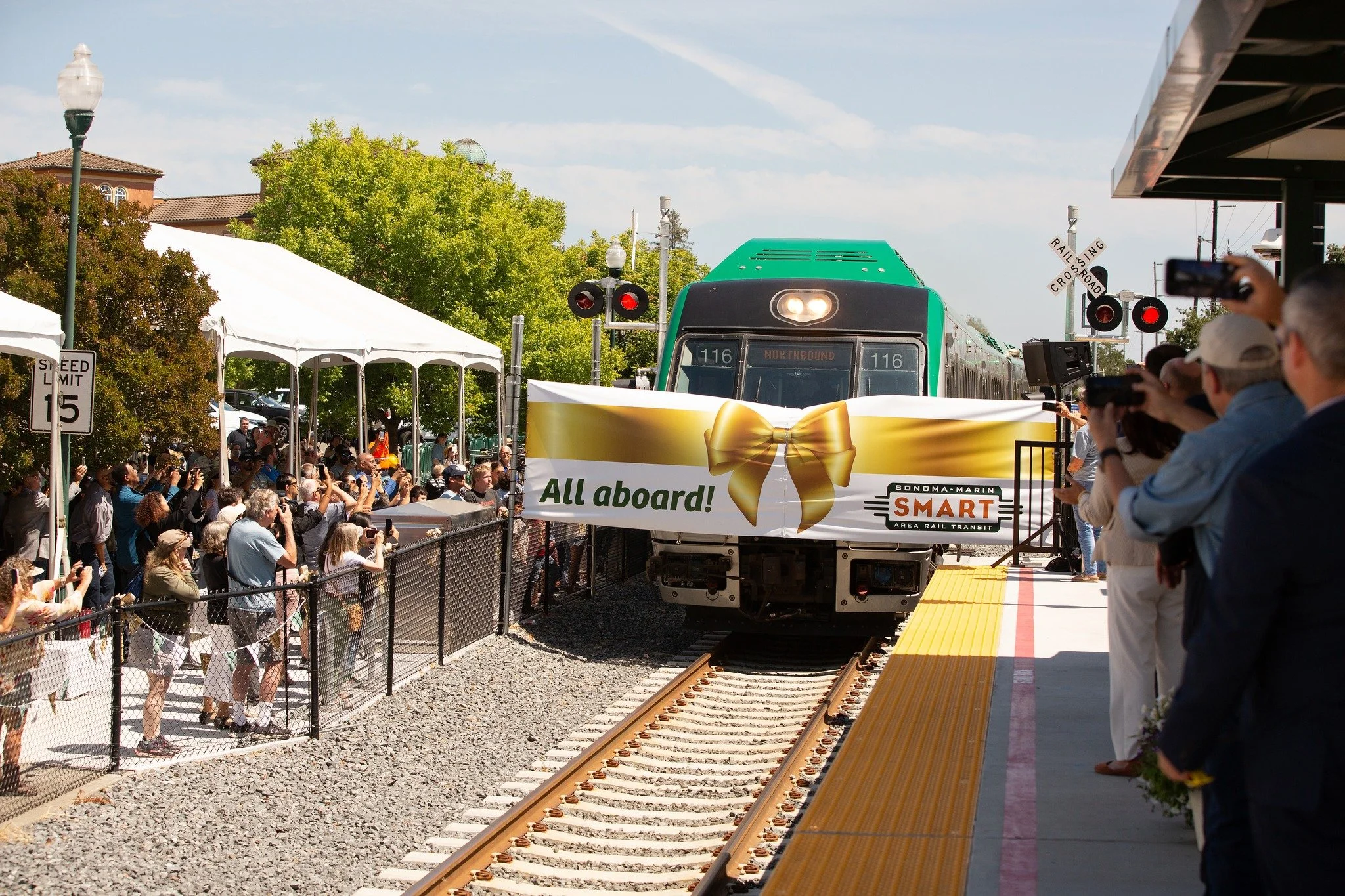 A train arriving at a station with a large ribbon and bow banner reading 'All aboard! SMART' and a crowd of people taking photos and watching.