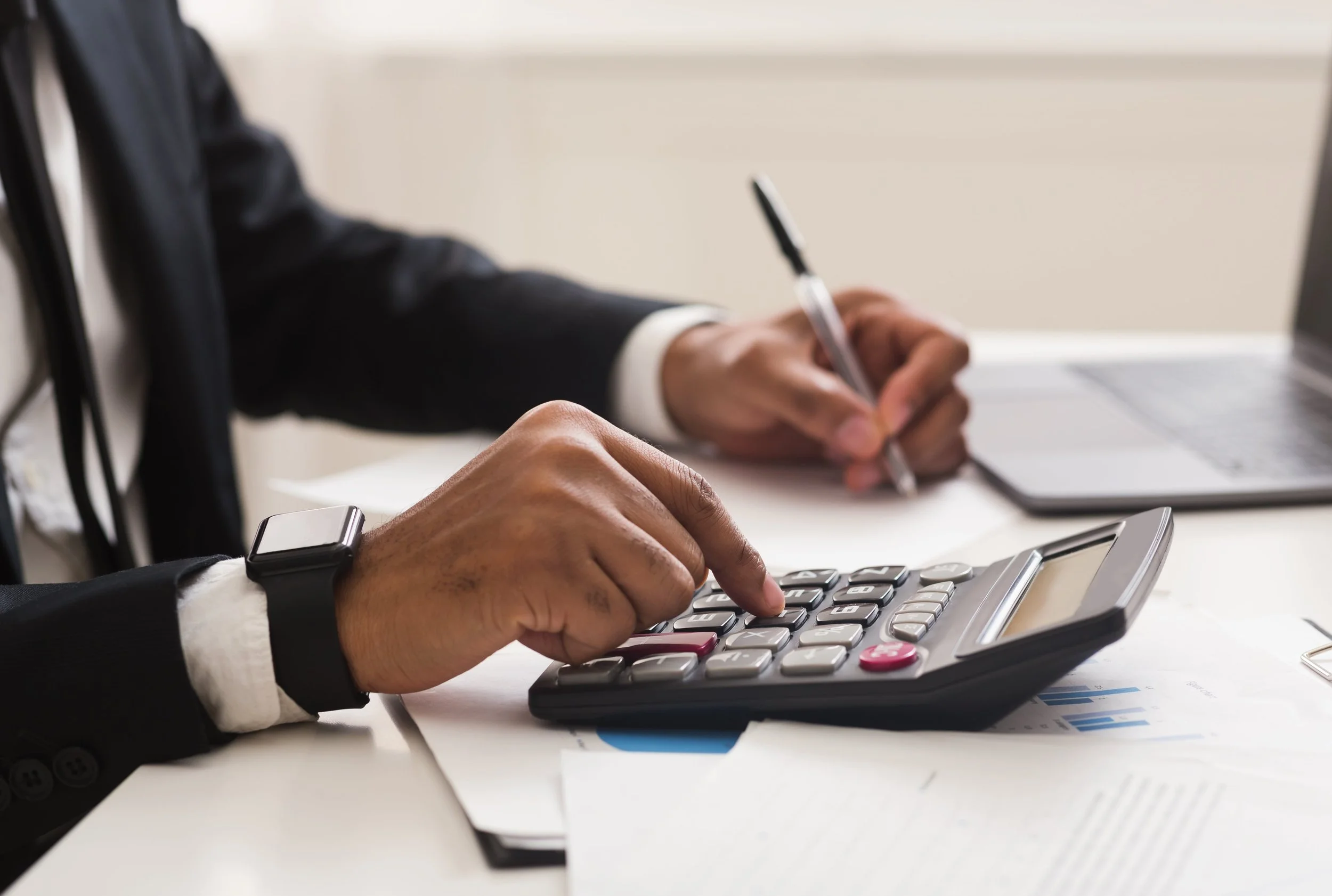 A person in a business suit using a calculator and writing on a notepad at a desk with documents and a laptop.