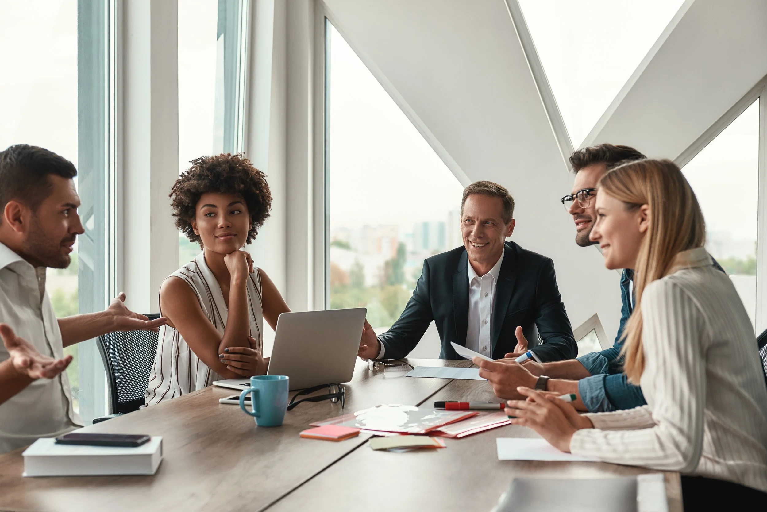 Five diverse professionals in a business meeting around a conference table, engaging in discussion and smiling, with large windows and cityscape in the background.