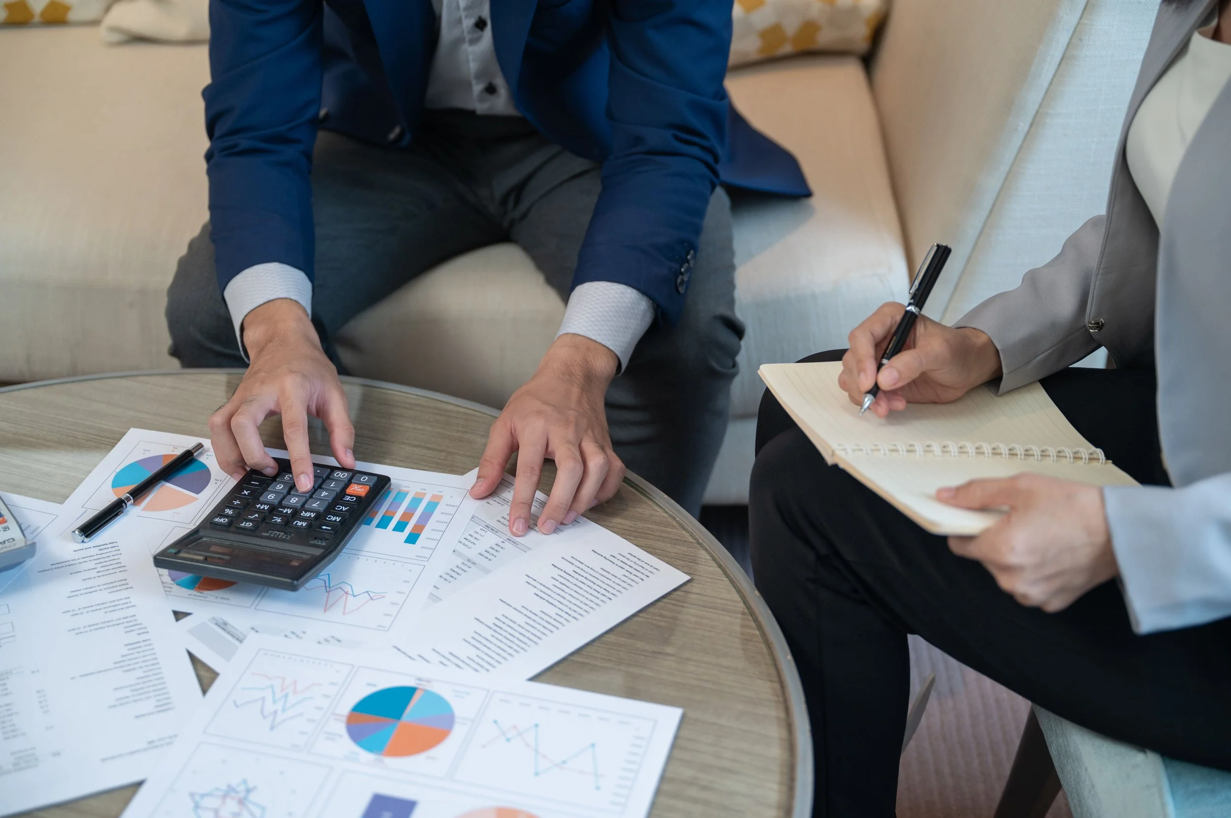 Two individuals conducting a business meeting with financial charts, a calculator, a pen, and documents spread on a round table.