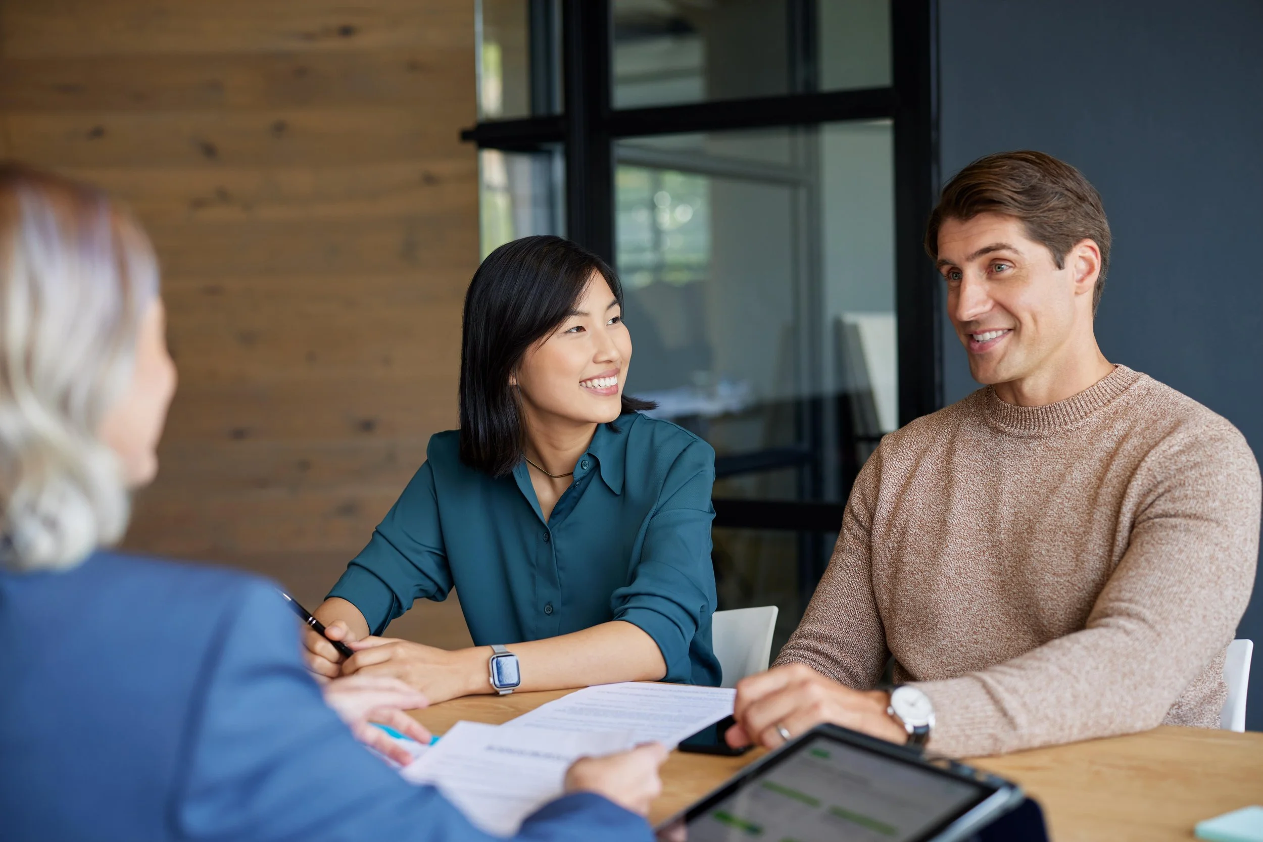 Three people having a meeting, two women and one man, sitting at a table with documents and laptops, in a modern office with wooden and glass walls.