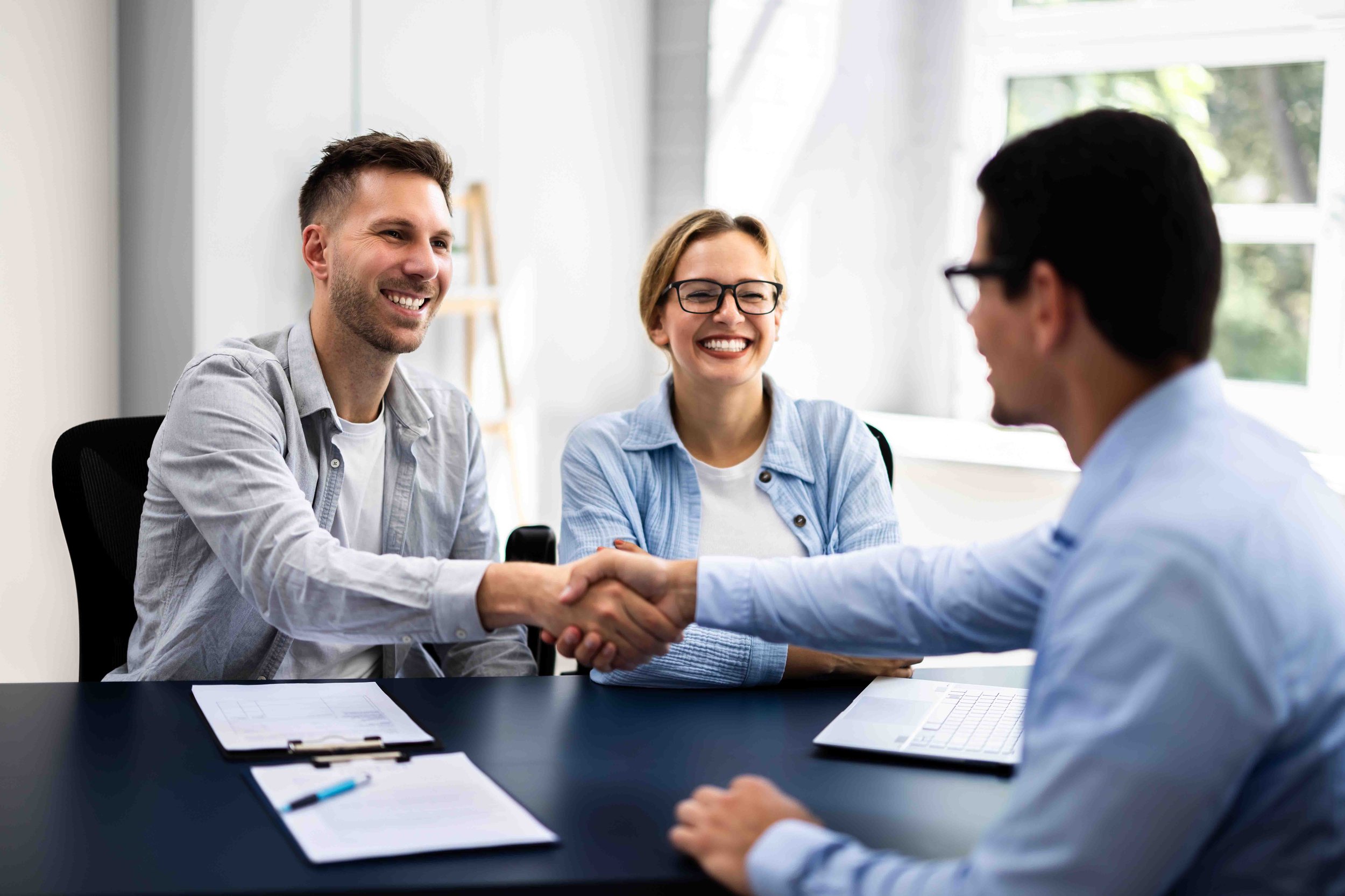 Three people in a meeting room, two men and one woman, smiling and shaking hands. The woman and the man on the left are sitting at a table with notebooks and a laptop in front of them, and the man on the right is facing them.