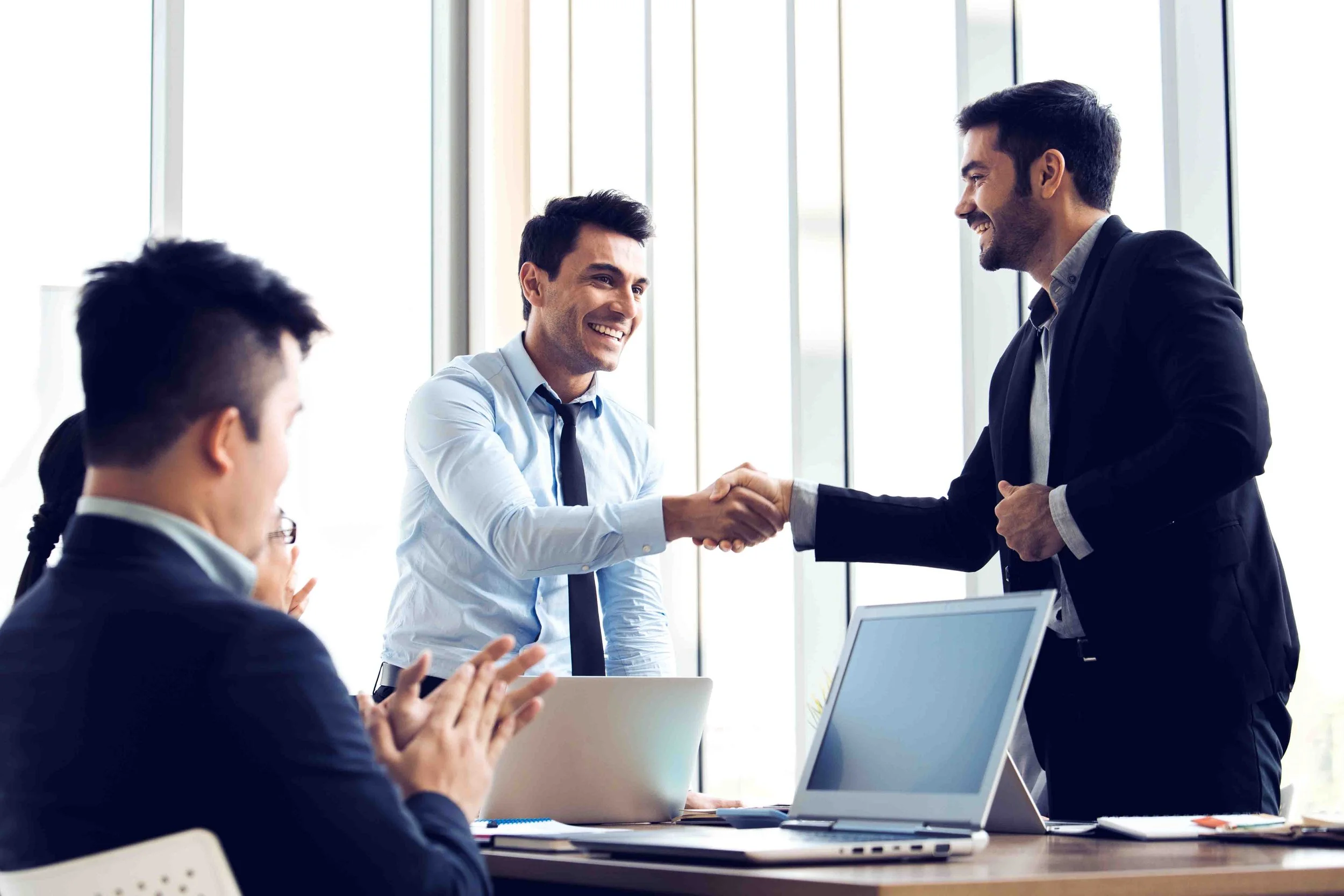 Two men shaking hands in a business setting, with two other people clapping and smiling.