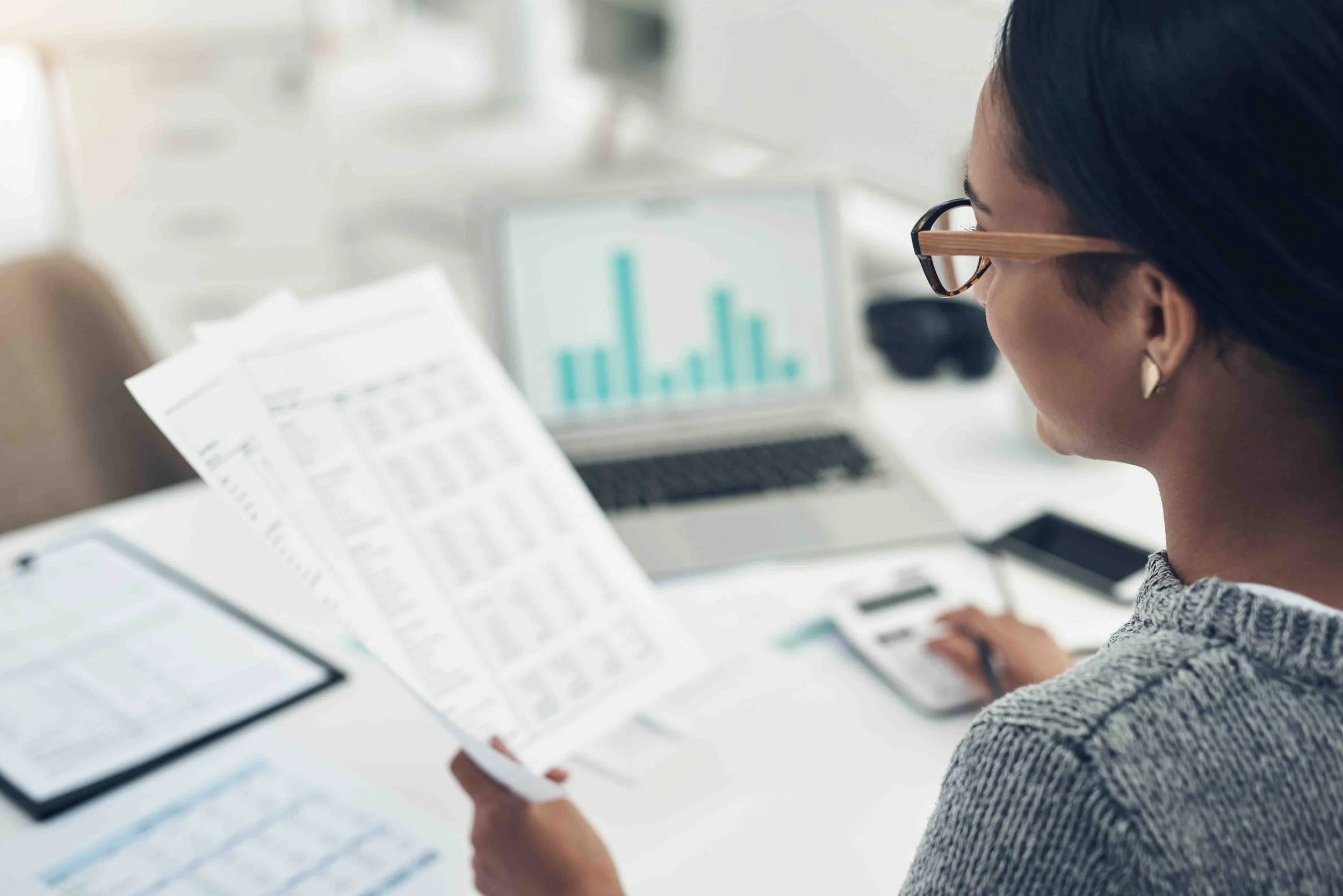 Woman reviewing financial documents at a desk with a laptop displaying a bar chart.
