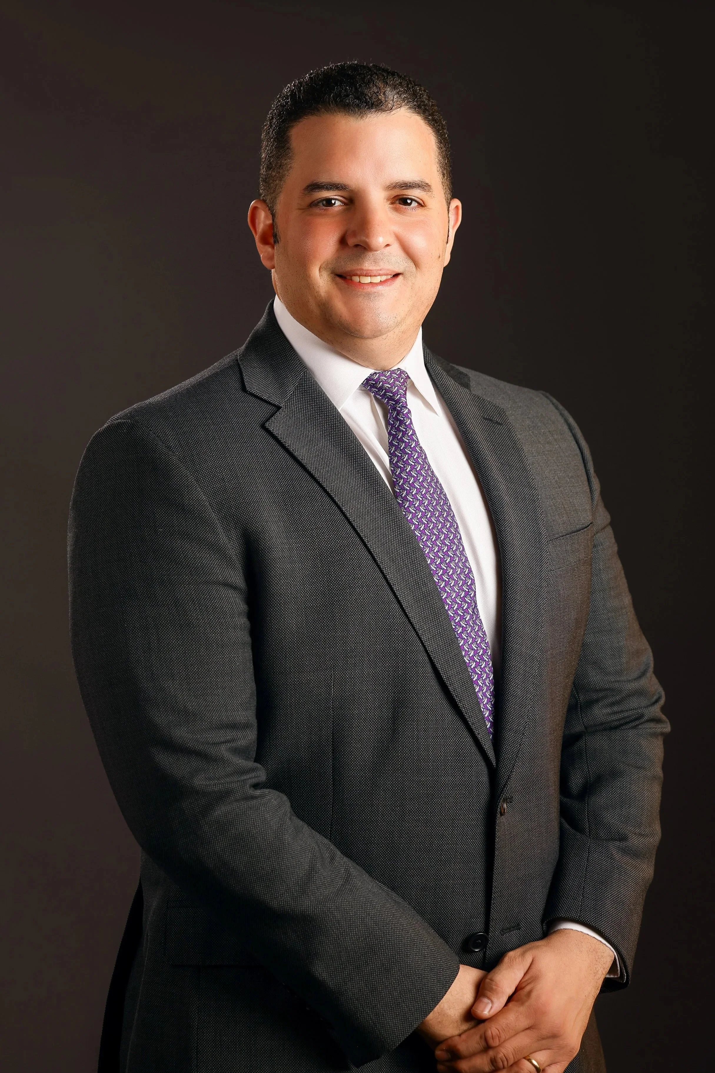 A professional man in a gray suit with a white shirt and purple patterned tie, standing with hands clasped in front of him, smiling at the camera against a dark background.