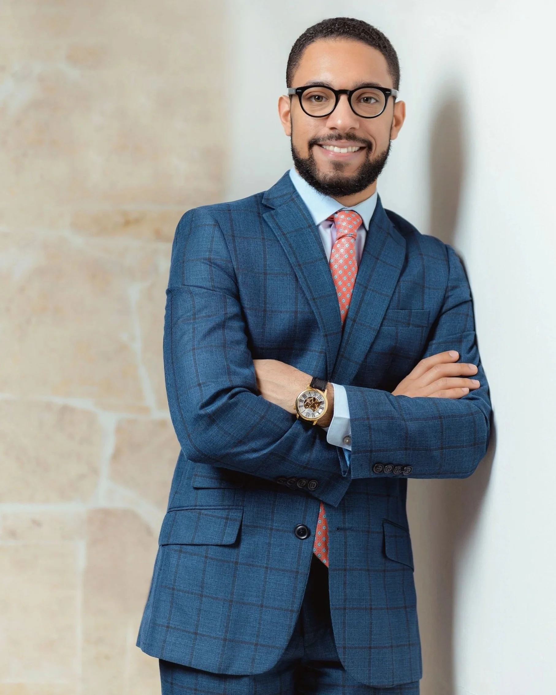 A man in a blue plaid business suit with glasses, a watch, and a pink tie stands with his arms crossed, smiling, against a brick and white wall.