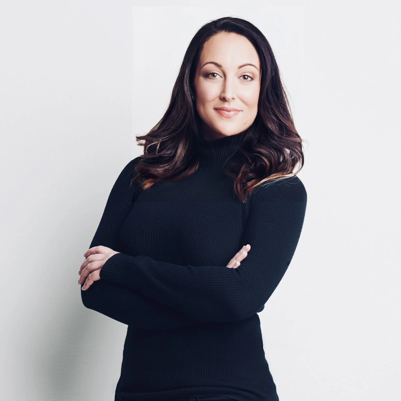 A woman with long dark hair standing against a plain white background, wearing a black turtleneck sweater and crossing her arms.