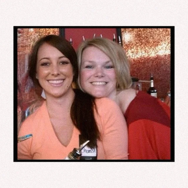 Two smiling women posing together indoors, one with dark brown hair and the other with blonde hair, both wearing colorful tops, with a warm, textured background.