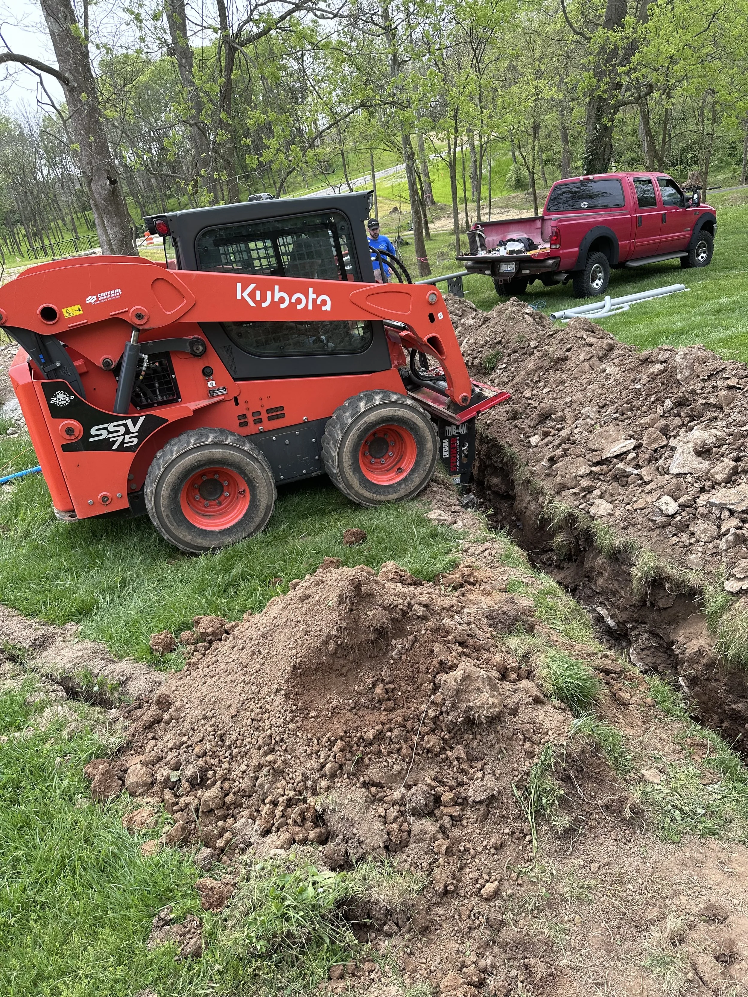 A Kubota compact excavator digging a trench in a grassy yard with a red truck parked in the background and trees surrounding the area.