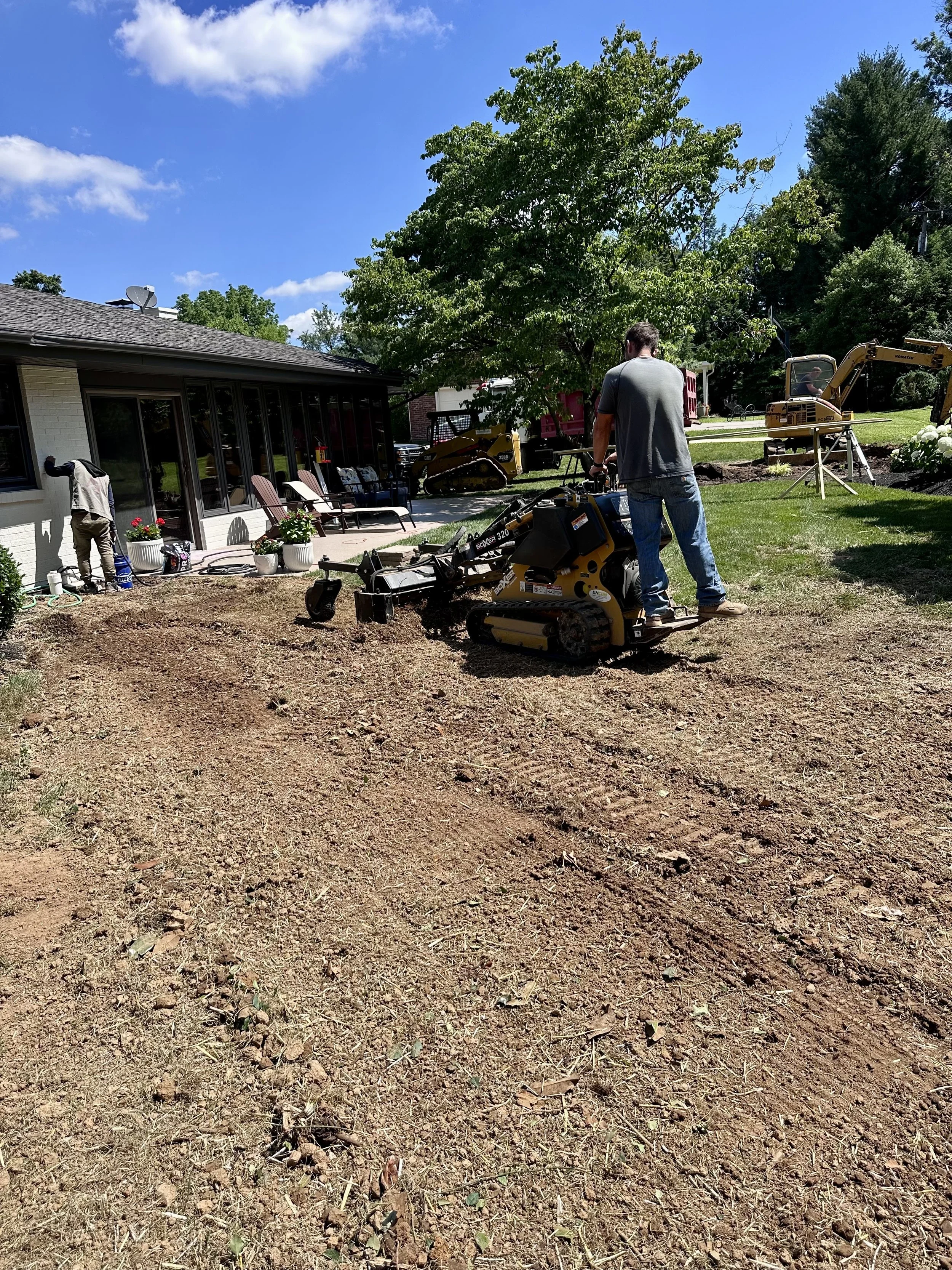 Two men working on landscaping a backyard, one using a mini roller and the other with a shovel, with trees, a house, and construction equipment in the background.