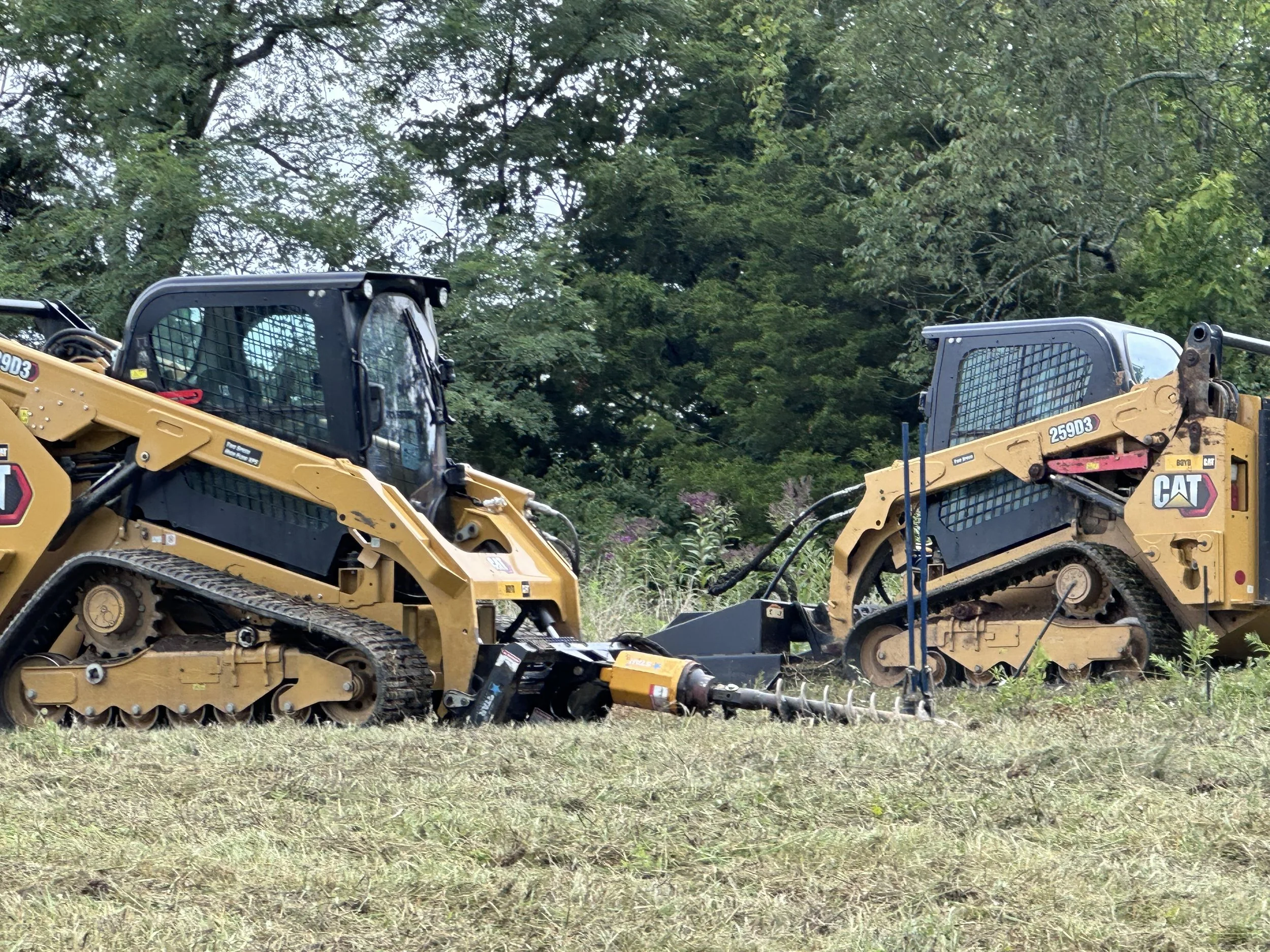 Two yellow Caterpillar tracked skid steer loaders on grassy field with trees in the background