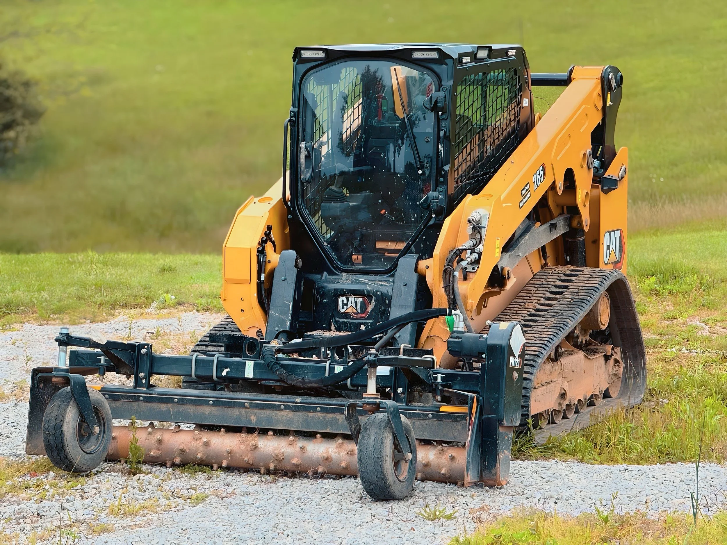 Yellow CAT skid steer loader on gravel with green grass and trees in the background.