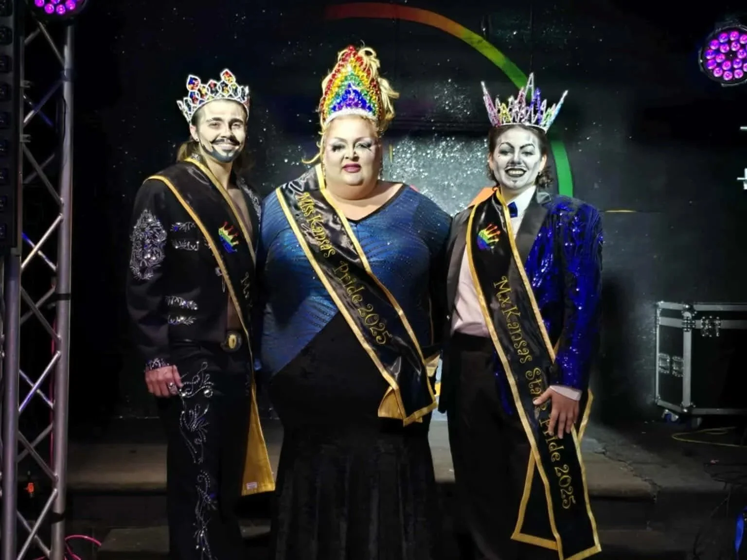Three individuals wearing crowns and sashes with the text 'Miss' and the year '2025,' dressed in elegant outfits, standing on a stage with colorful lighting.
