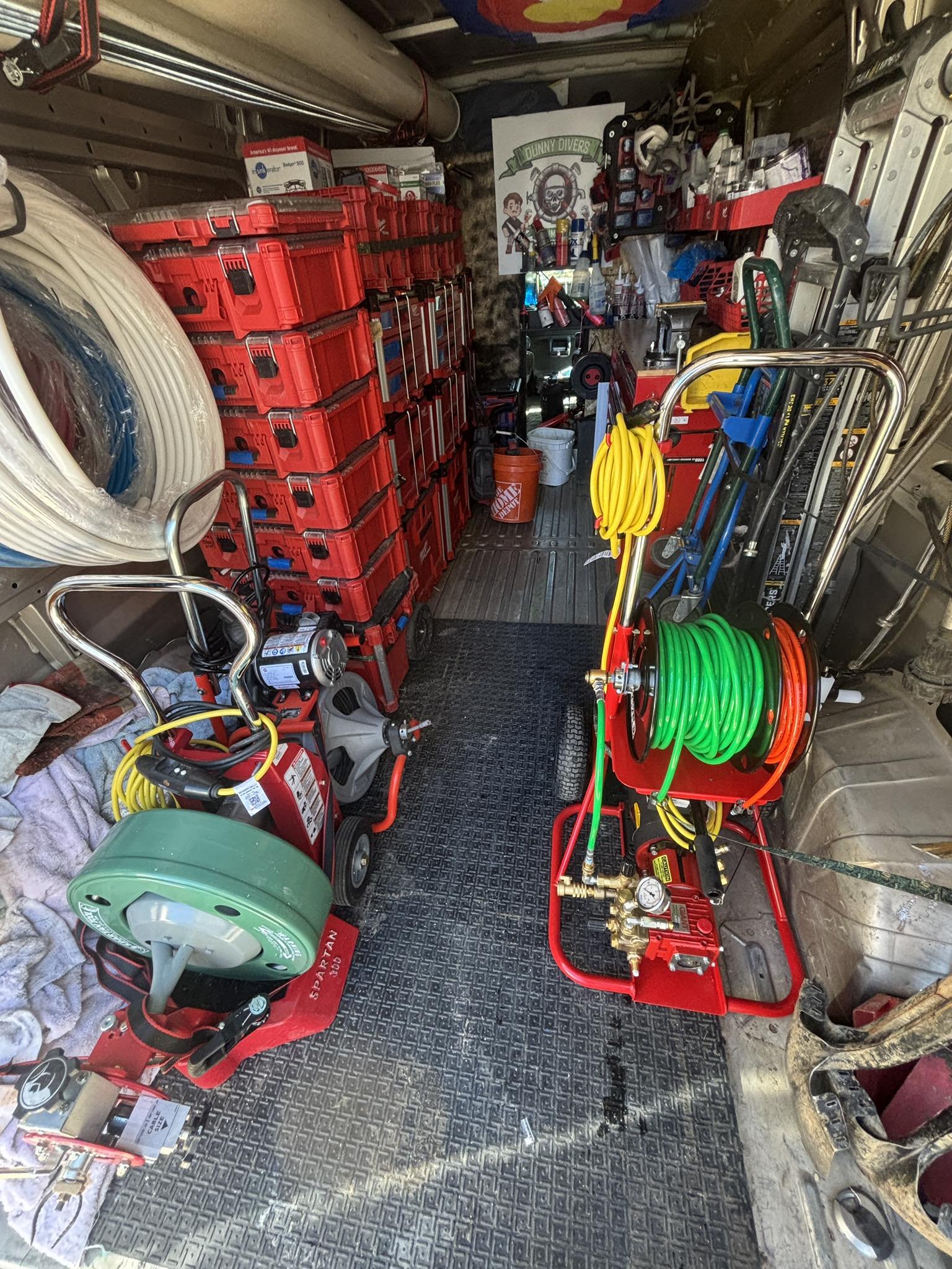 The inside of a utility vehicle or work truck cluttered with equipment, tools, and supplies, including red storage containers, garden hoses, and various maintenance and cleaning gear.