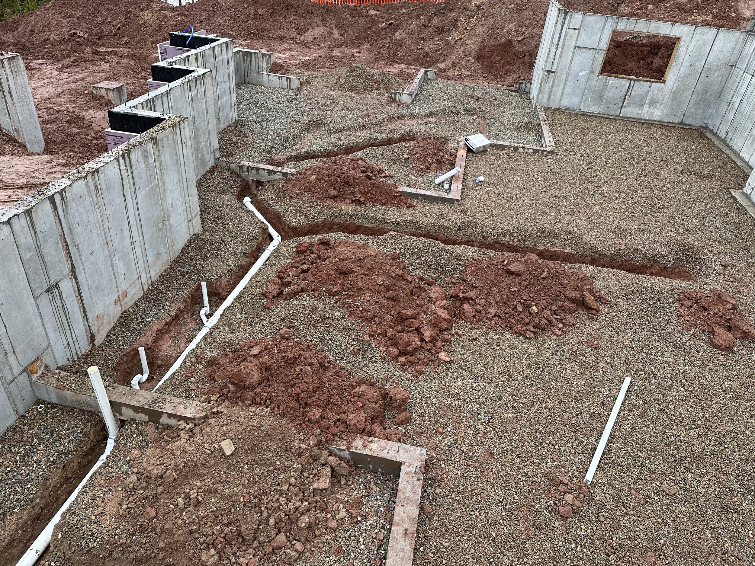 Construction site foundation with concrete walls and exposed pipes, dirt and gravel ground.
