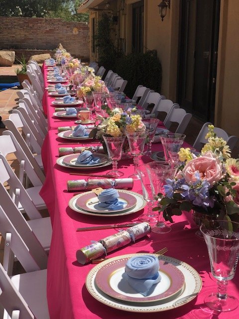 A long outdoor table setting (tablescape) for a celebration with pink tablecloth, floral centerpieces, plates with blue napkins, and glassware.
