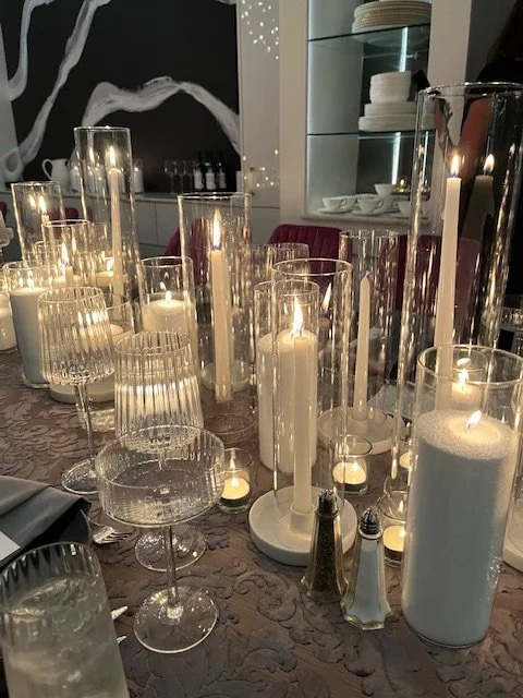 A dining table decorated with various tall white sand candles in glass holders, creating a cozy atmosphere.