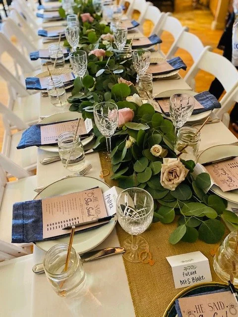 A long wedding reception table decorated with a green foliage and pink rose floral centerpiece, white plates, silverware, cloth napkins, and glassware, with personalized name cards.