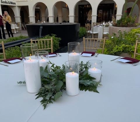 Round table set for a formal event with white tablecloth, five candles in glass holders, and burgundy napkins, surrounded by gold chairs.